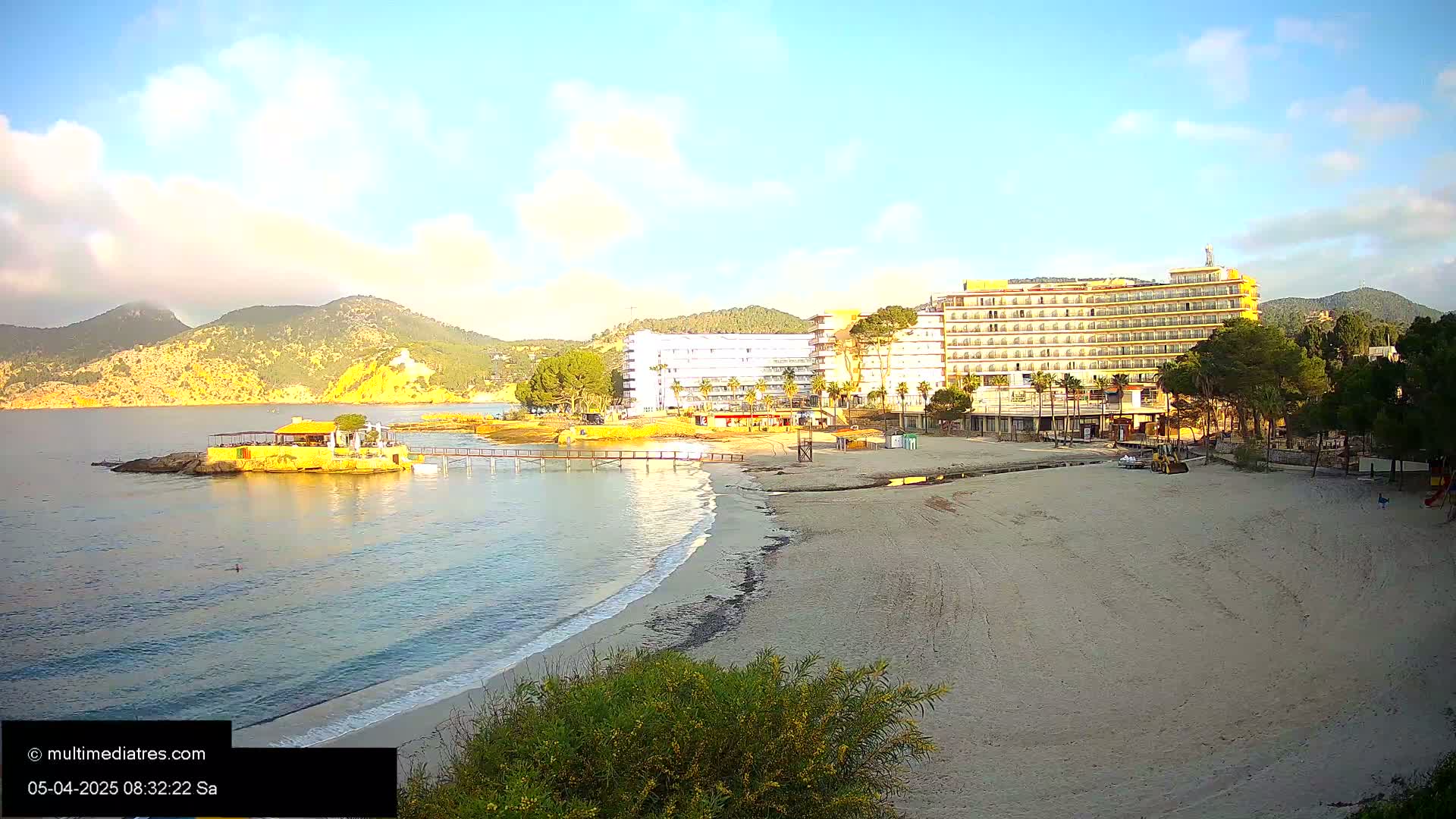 A sandy beach with calm, clear water laps gently at the shore, next to a large hotel and a small restaurant on a rock outcropping, all under a partly cloudy, sunny sky.