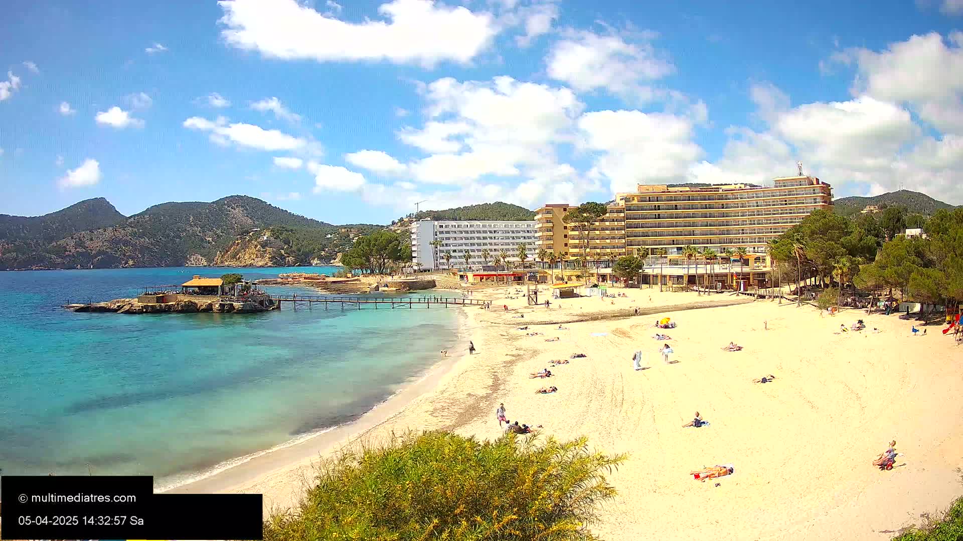 A sandy beach with sunbathers stretches out below a large hotel, a small pier leading to a beachfront restaurant, all under a partly cloudy sunny sky, with mountains visible in the background.
