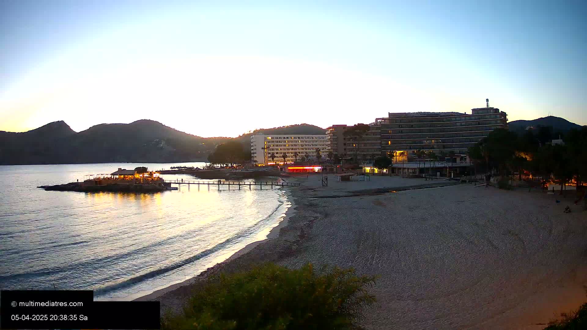 A sandy beach at sunset, with calm waves lapping the shore, is bordered by a seaside restaurant, hotels, and vegetation, all nestled against a backdrop of hills.