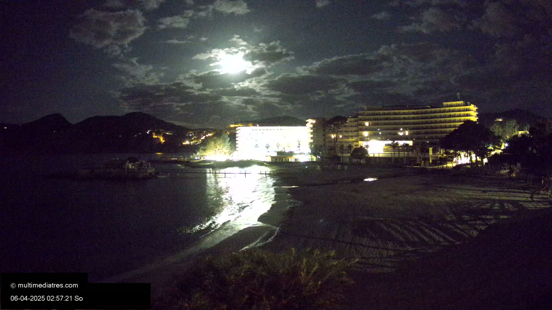 A moonlit night scene shows a brightly lit hotel on a beach near dark mountains, with calm water reflecting the lights.