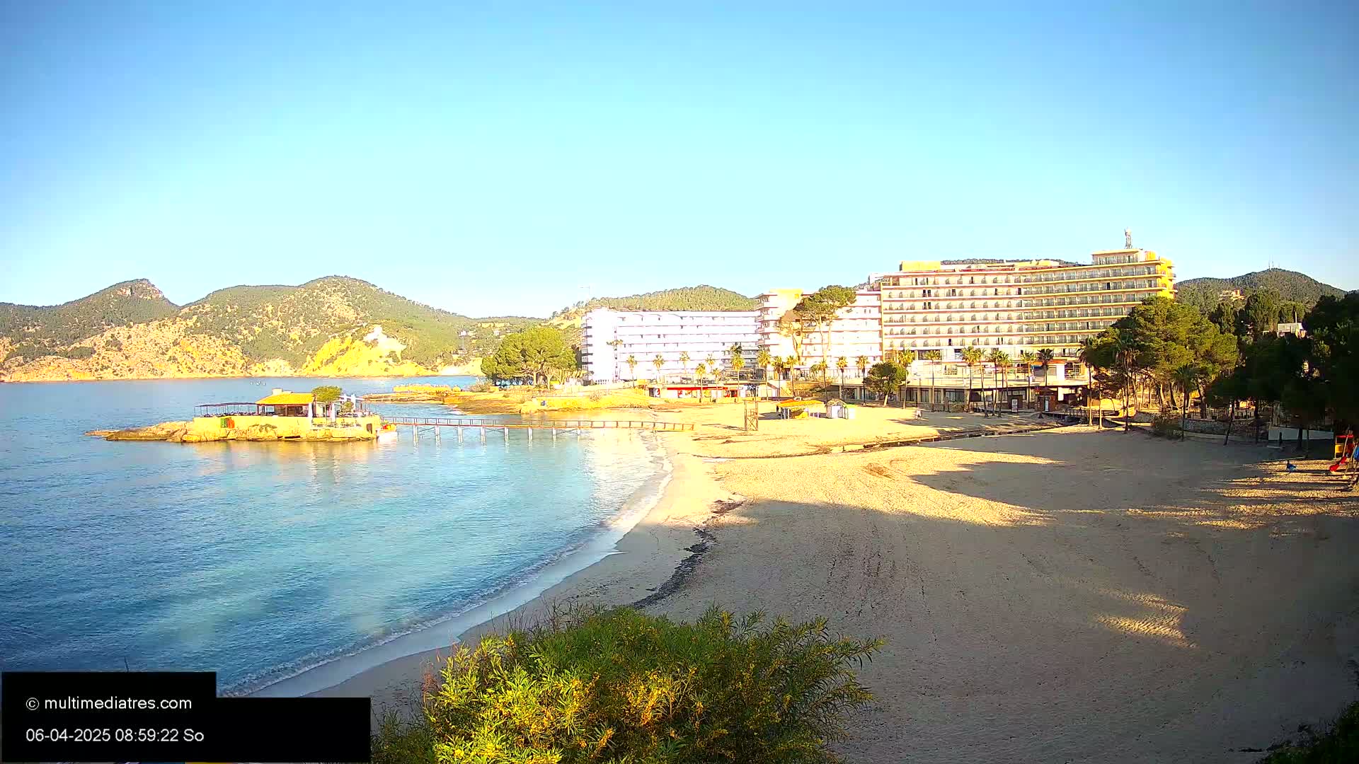A sandy beach with clear blue water, a small restaurant on a pier, and a large hotel in the background under a bright sunny sky.