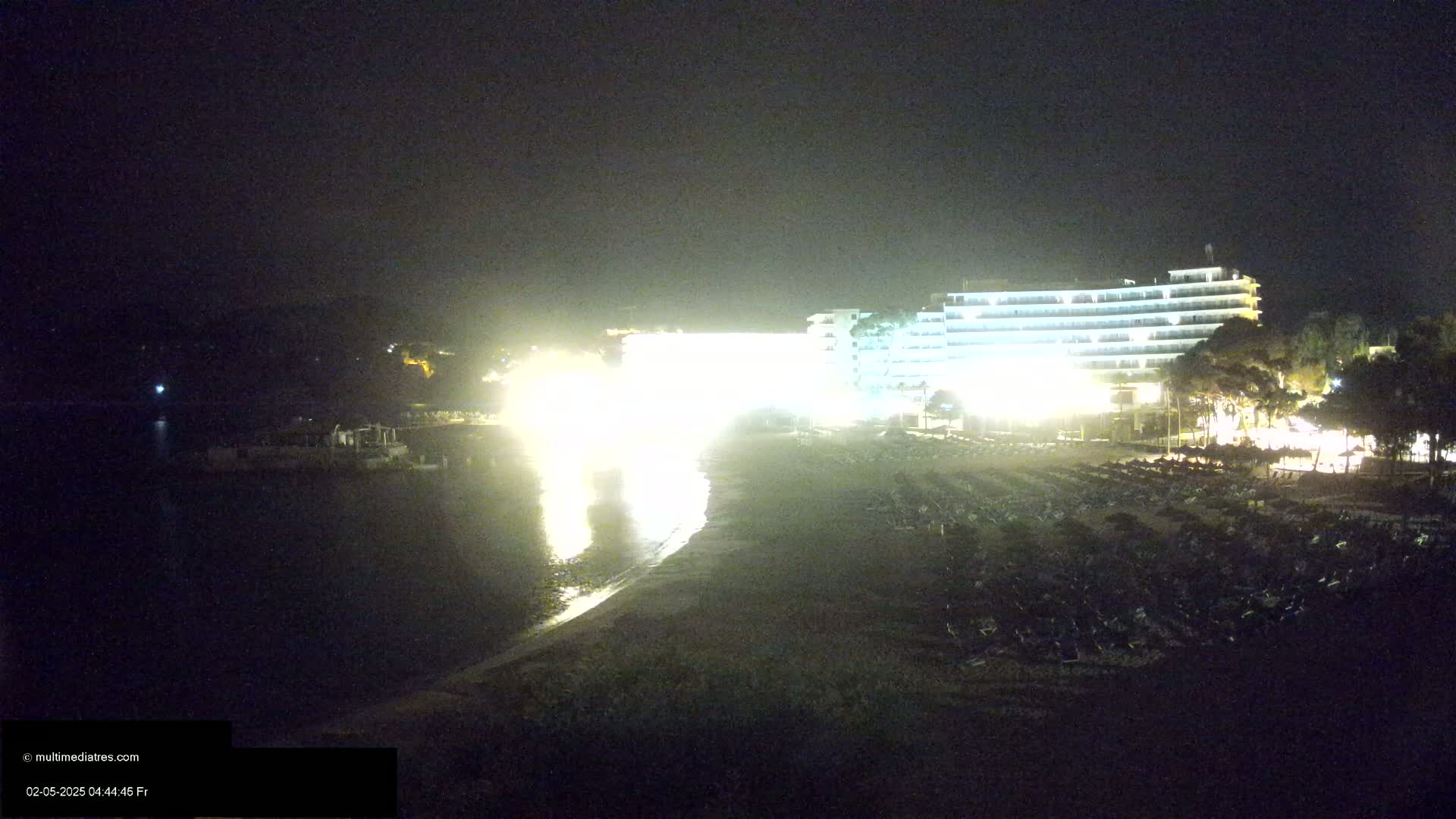 A brightly lit hotel stands on a dark beach at night next to calm water.