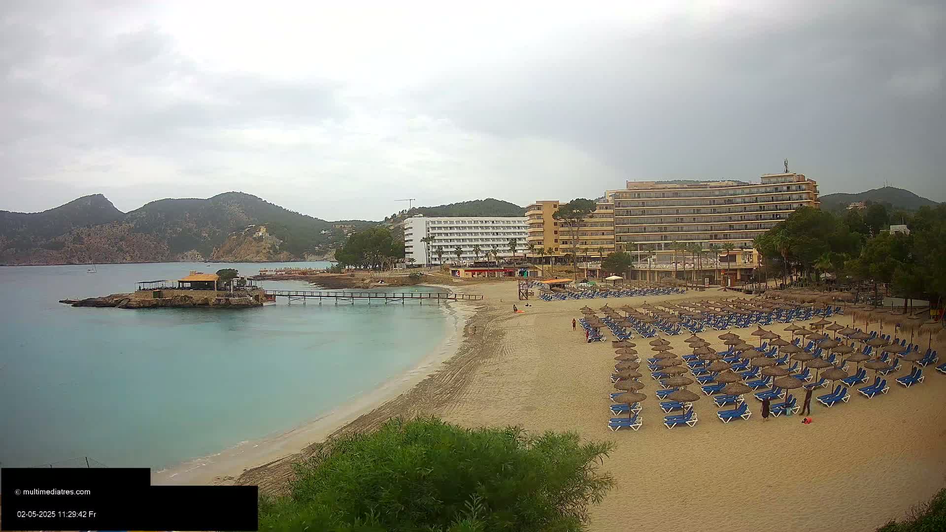 A sandy beach with rows of blue lounge chairs and thatched umbrellas sits in front of several hotels, under an overcast sky, with calm, turquoise water and mountains in the background.
