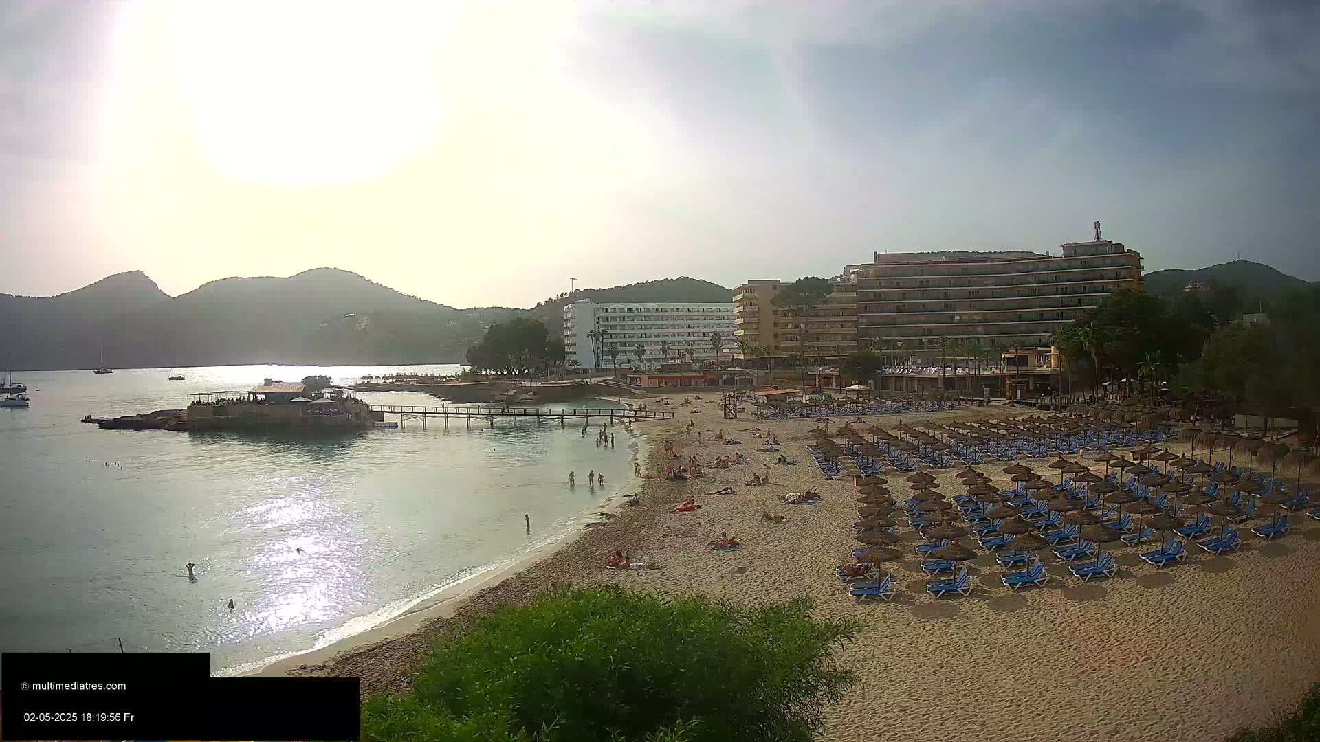 A sandy beach with numerous blue lounge chairs and thatched umbrellas is situated in front of several hotels and a calm bay under a partly cloudy sky.