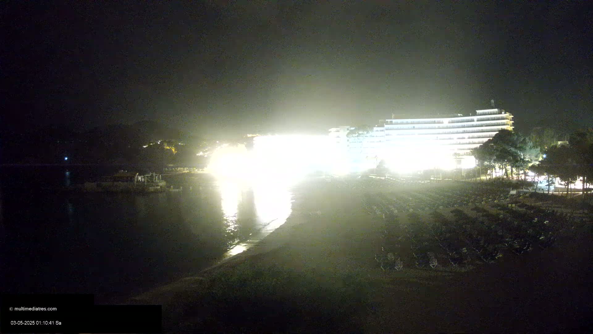 A brightly lit hotel overlooks a dark beach at night with many chairs set up on the sand, under a hazy sky.
