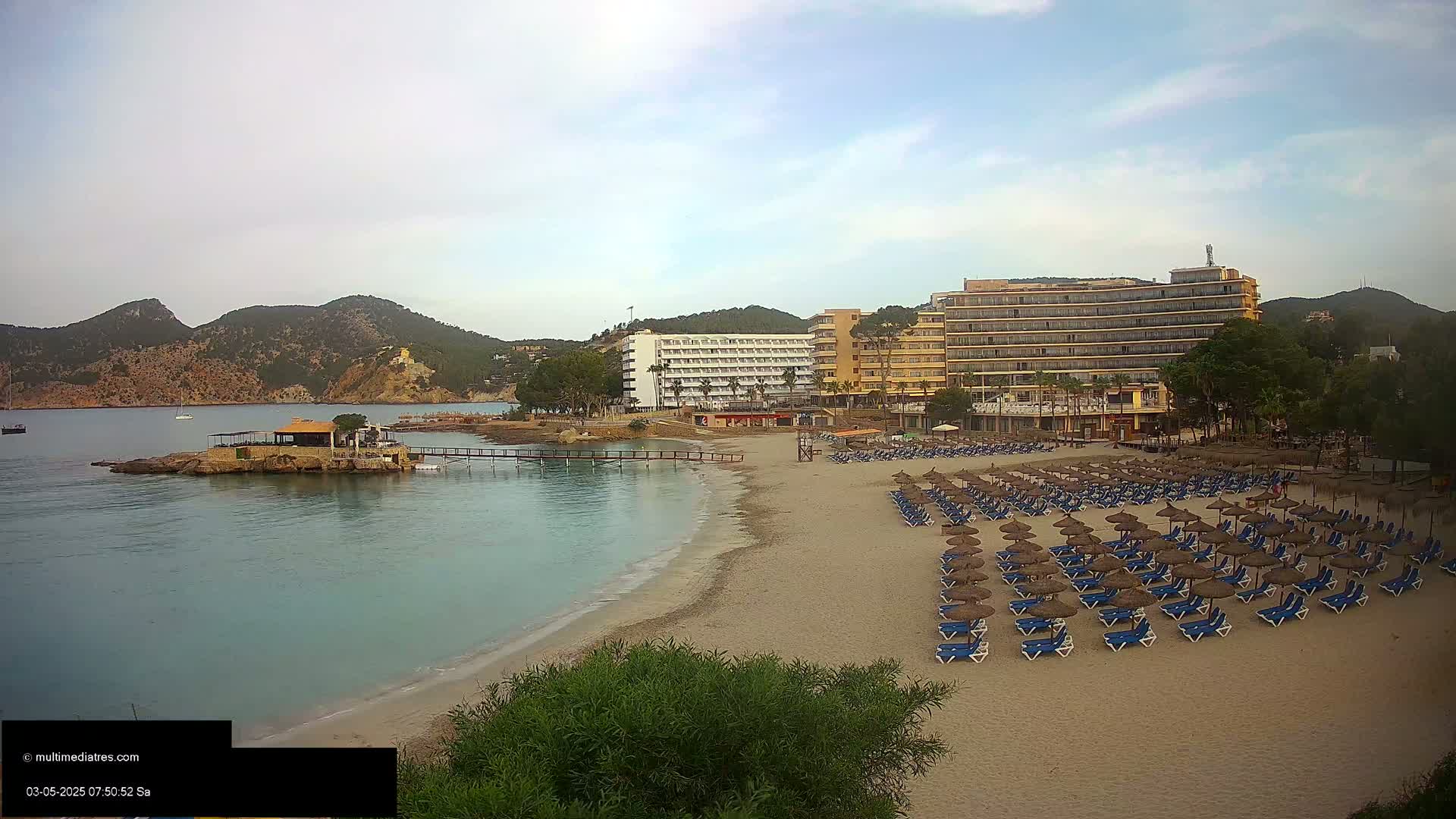 A sandy beach with rows of unoccupied lounge chairs and umbrellas sits in front of several hotels and a small island with a building, all under a mostly clear sky.