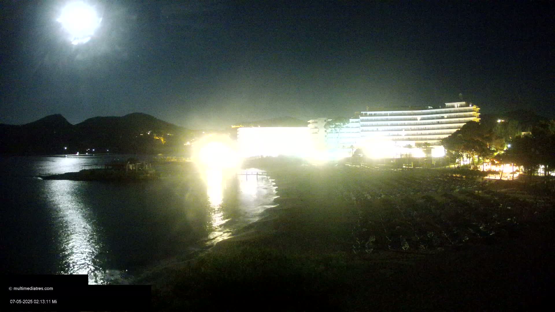 A brightly lit hotel stands on a beach at night under a full moon, with dark mountains in the background.