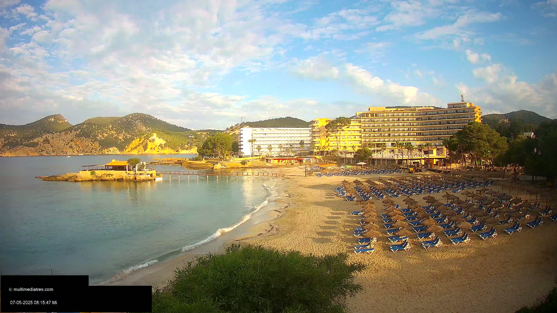 A sandy beach with rows of lounge chairs and umbrellas sits in front of several hotels, with calm, clear water lapping at the shore under a partly cloudy, sunny sky, backed by low mountains.