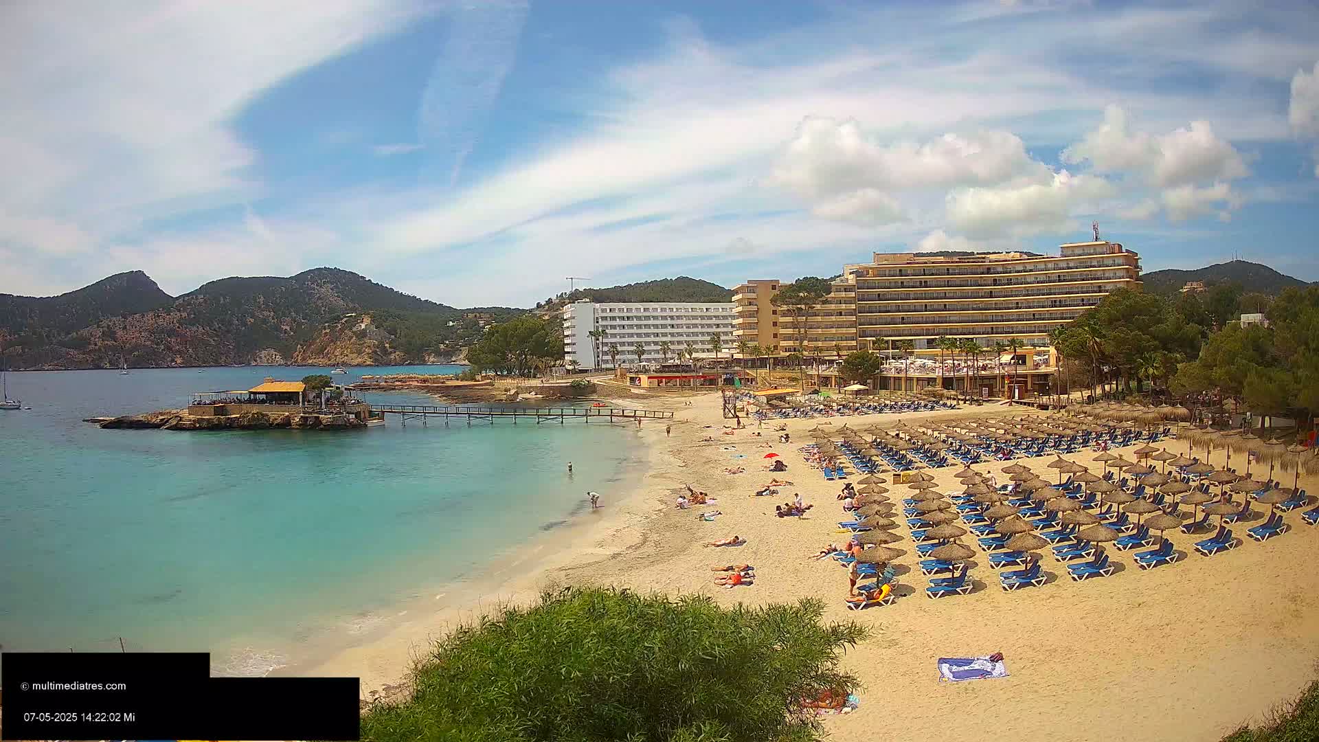 A sandy beach with numerous blue lounge chairs and thatched umbrellas stretches out before a large hotel complex, all under a partly sunny sky.