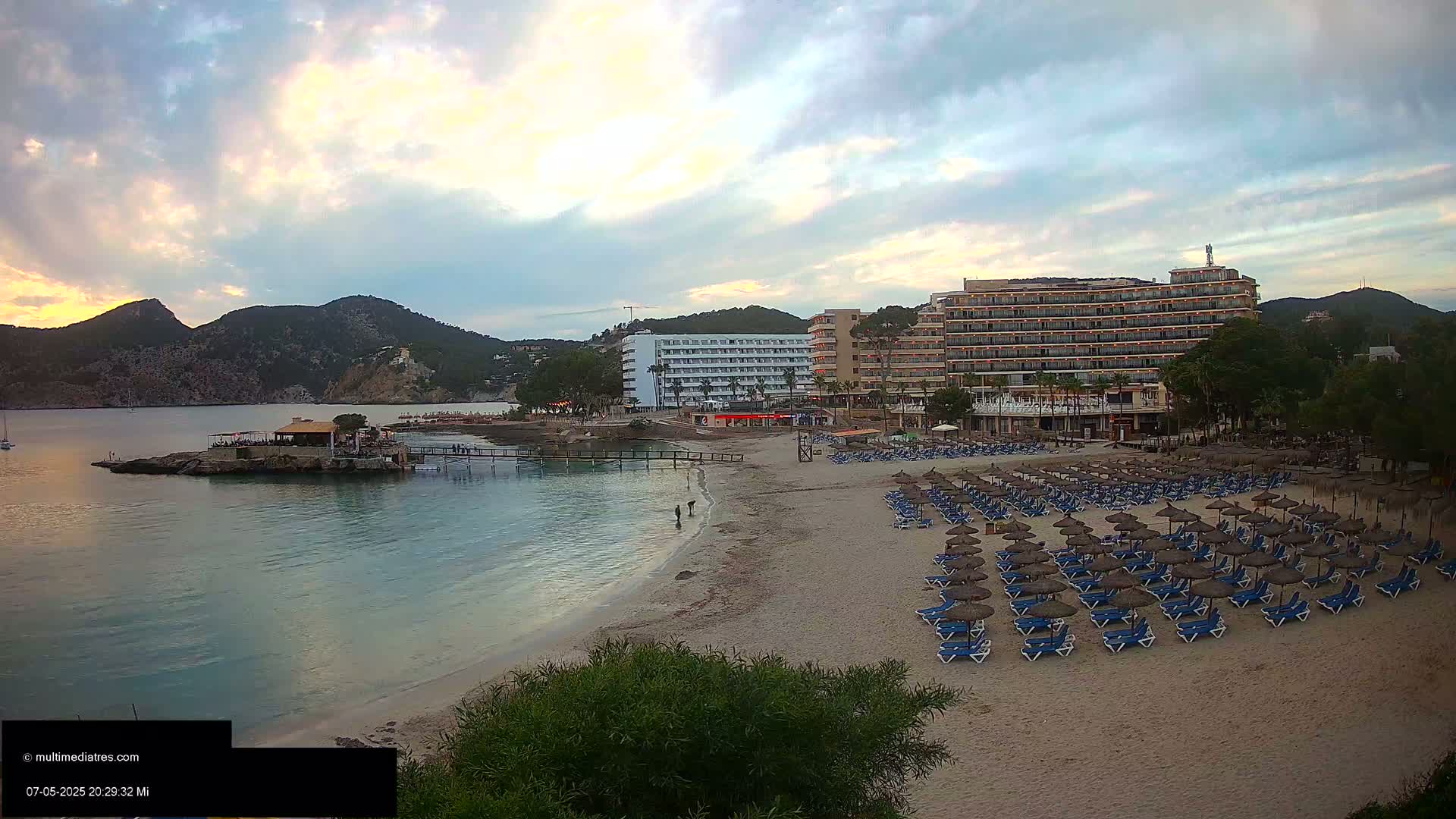 A sandy beach with numerous lounge chairs and umbrellas sits below two large hotels,  under a partly cloudy sunset sky, next to calm, clear water and a rocky shoreline.