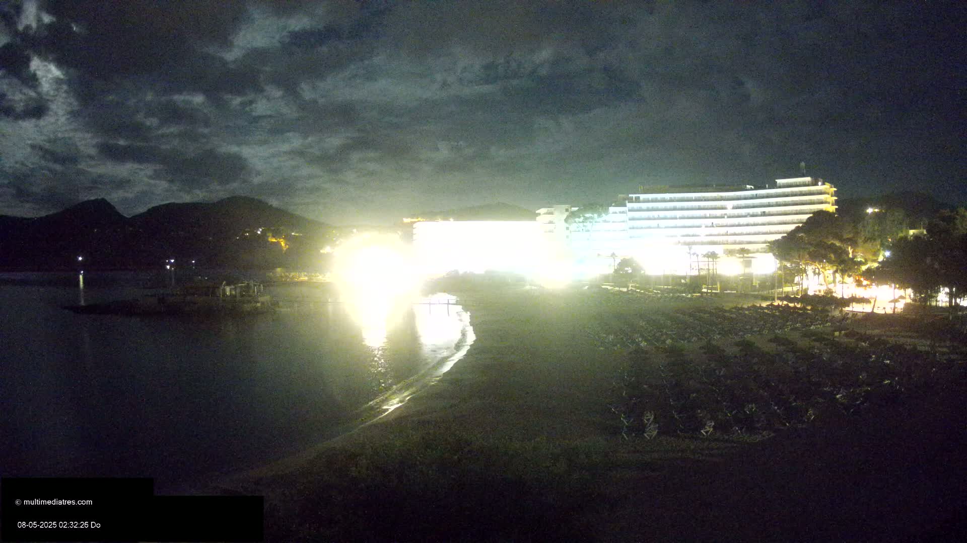 A brightly lit hotel sits on a beach at night under a cloudy sky, with calm water reflecting the lights.