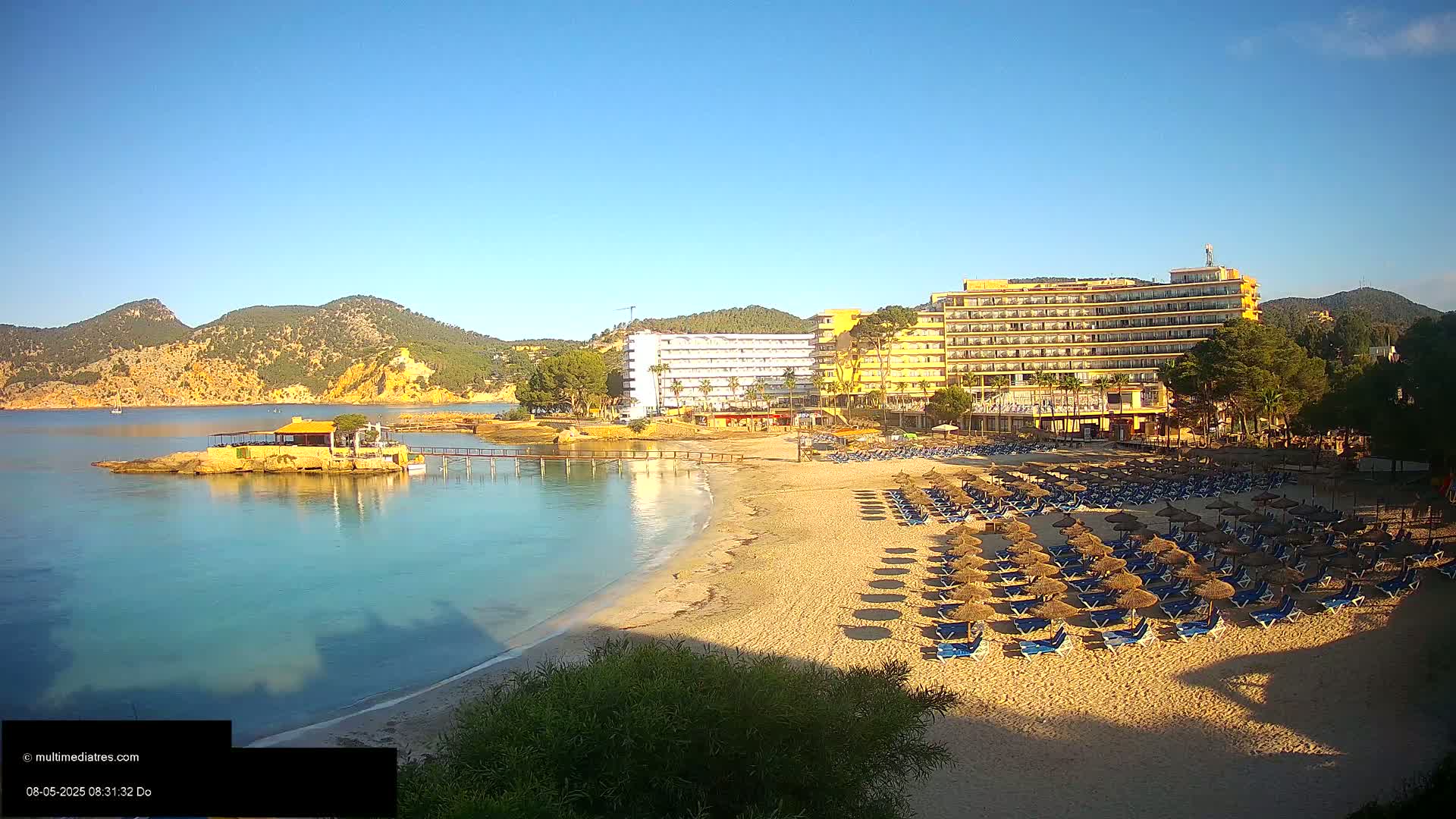 A sandy beach with numerous umbrellas and lounge chairs sits before a large hotel complex and calm turquoise water, all under a clear blue sky.