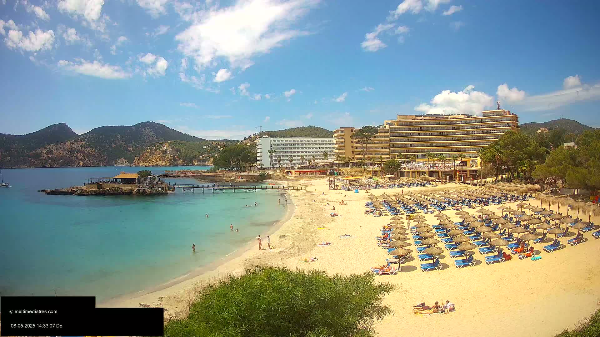A sandy beach with numerous lounge chairs and umbrellas, situated next to a large hotel and calm turquoise water under a partly sunny sky.