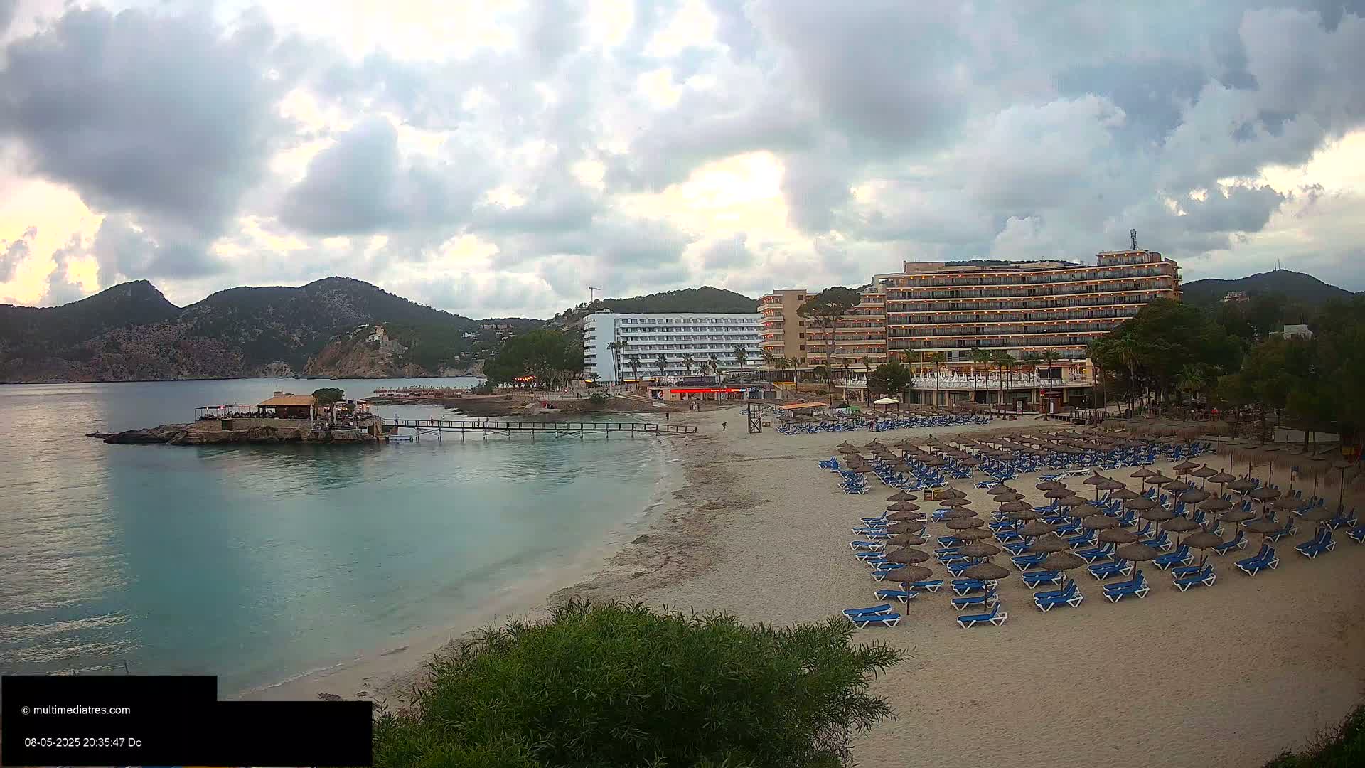 A sandy beach with numerous blue lounge chairs and thatched umbrellas sits below two hotels near calm, turquoise water, backed by hills under a partly cloudy sky.