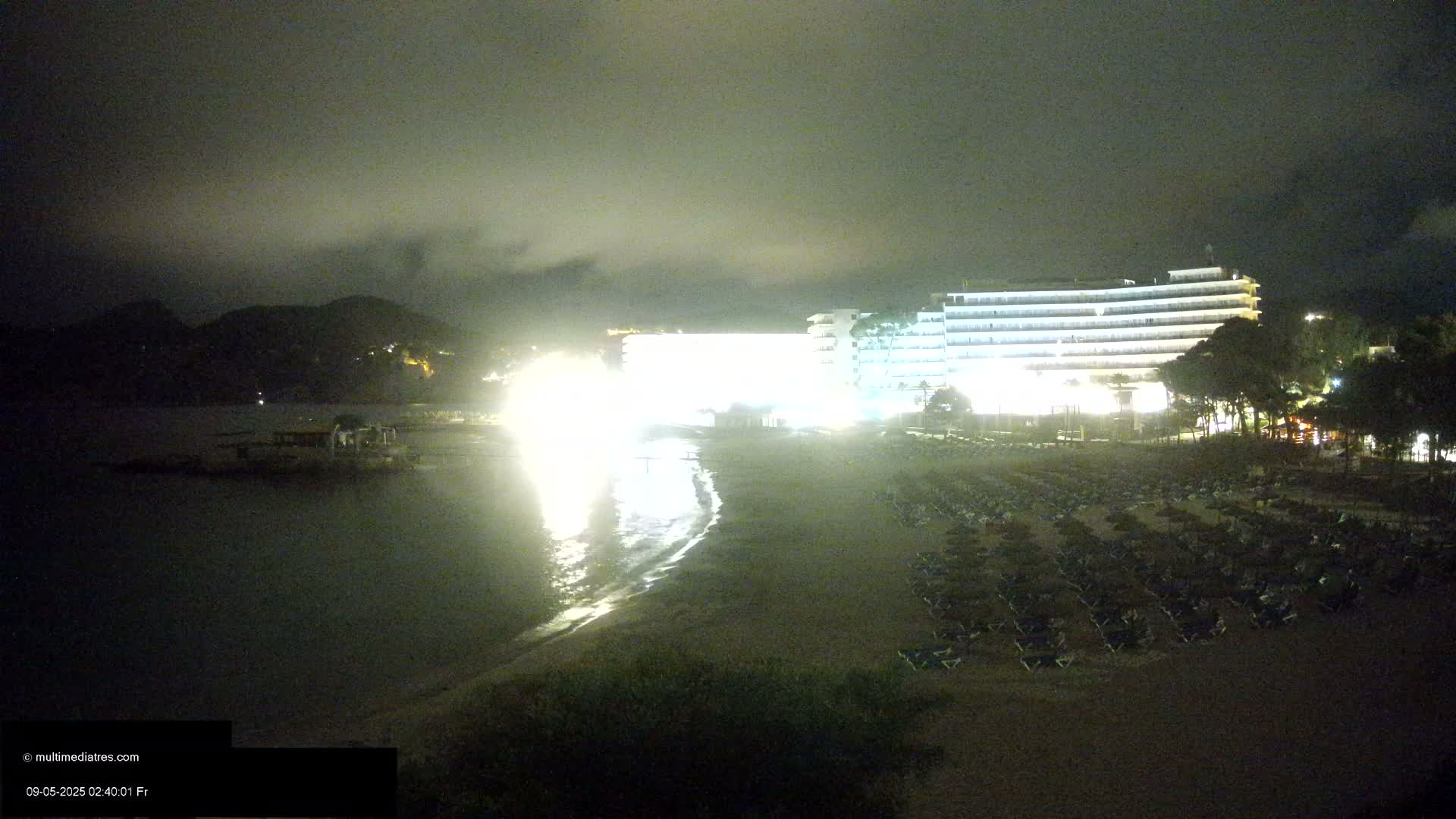 A brightly lit hotel stands on a beach at night, with rows of closed beach chairs and umbrellas in front of it, under a dark, cloudy sky.