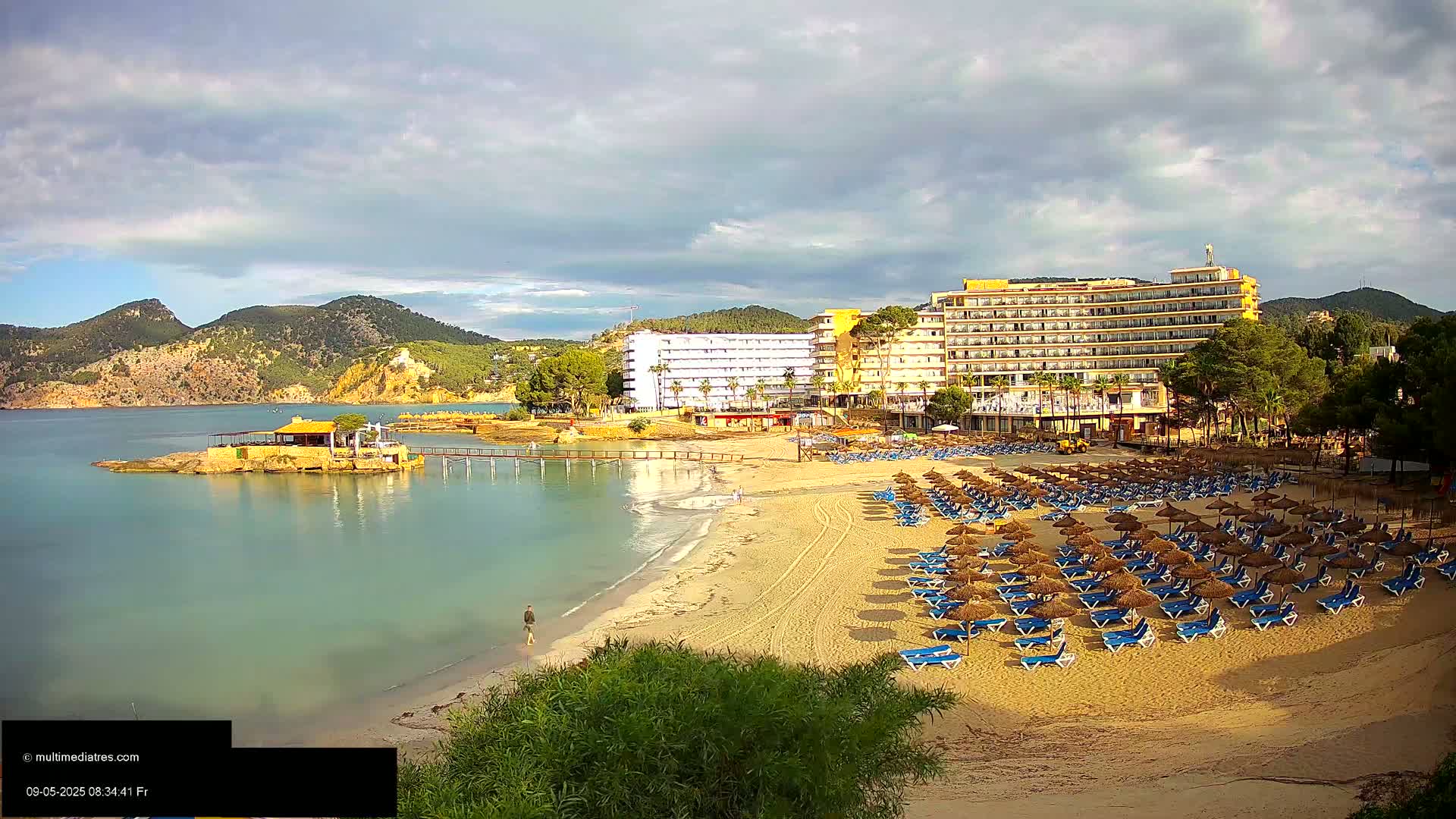 A sandy beach with numerous blue lounge chairs and straw umbrellas is situated in front of several large hotels, overlooking a calm bay and mountainous landscape under a partly cloudy sky.