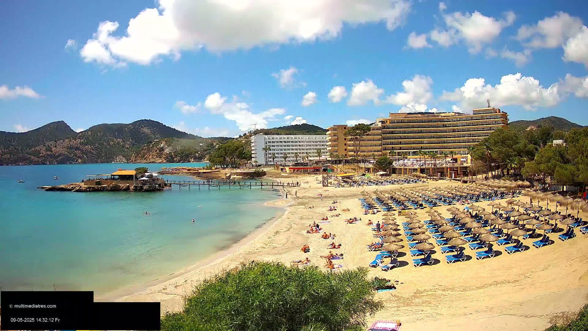 A sandy beach with numerous thatched umbrellas and lounge chairs is situated next to a calm, turquoise bay, with a hotel and green hills in the background under a partly sunny sky.