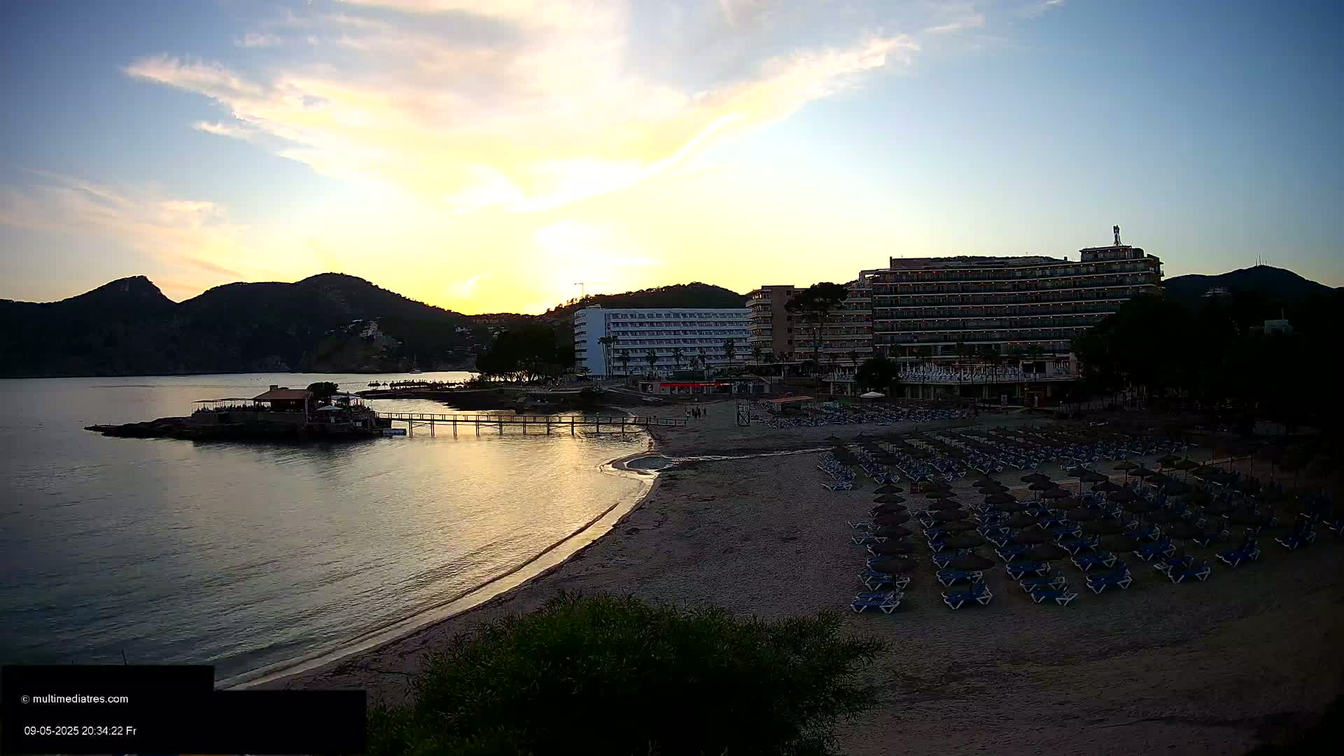 A sandy beach with rows of empty lounge chairs and umbrellas sits at the base of a hotel under a partly cloudy sunset sky near calm water and mountains.
