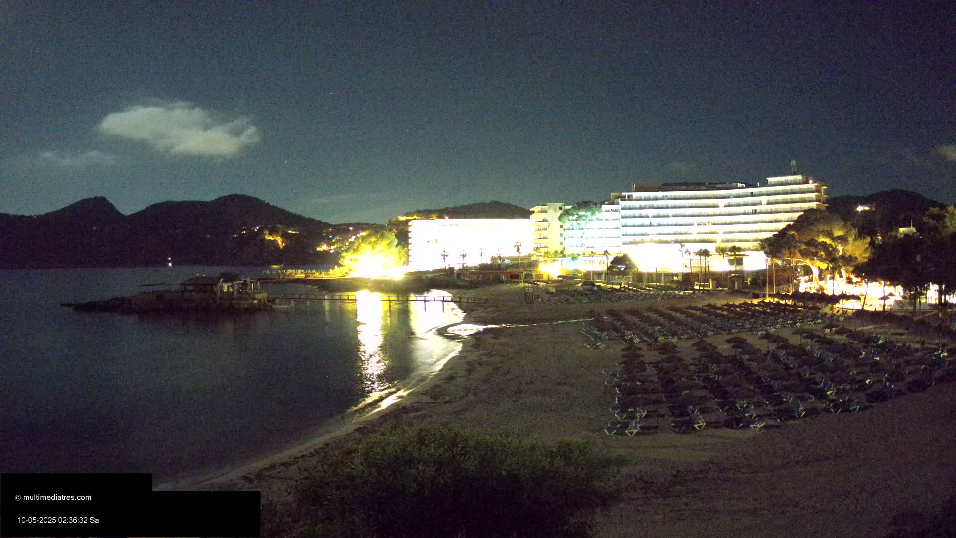 A nighttime view of a beach with many lounge chairs and umbrellas, a brightly lit hotel, and calm water under a partly cloudy sky.
