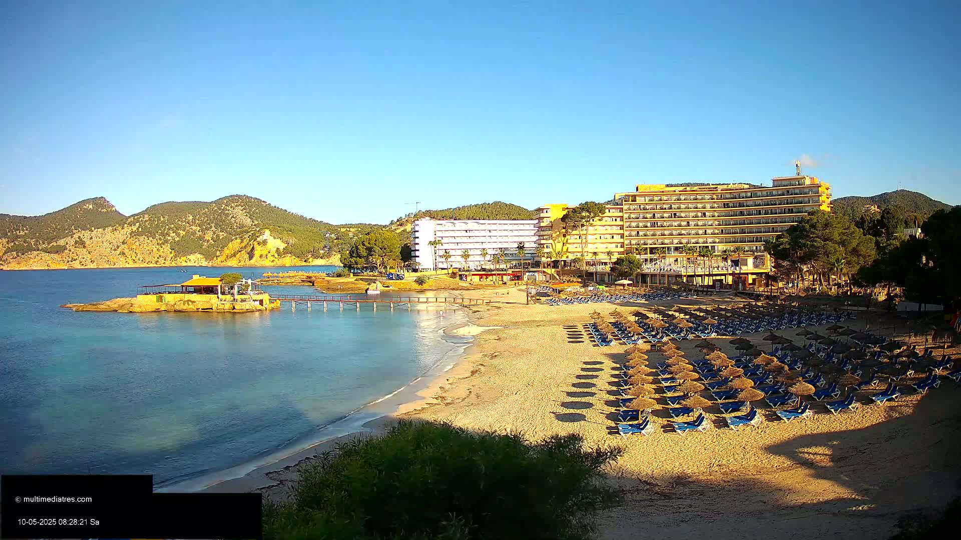 A sandy beach with numerous thatched umbrellas and lounge chairs sits below hotels and a small restaurant on a rocky outcropping, all under a clear blue sky.