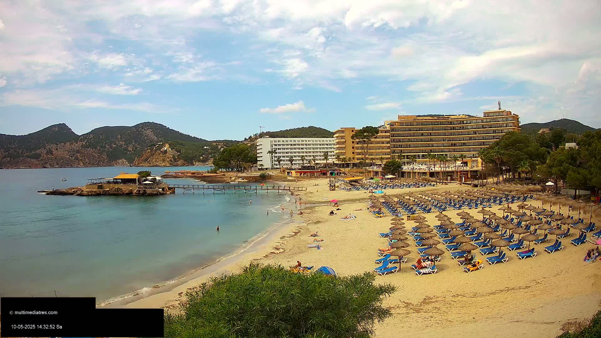A sandy beach with numerous lounge chairs and umbrellas under a partly cloudy sky, adjacent to several hotels and a small restaurant on a rocky outcropping, with calm, clear water in a bay surrounded by hills.