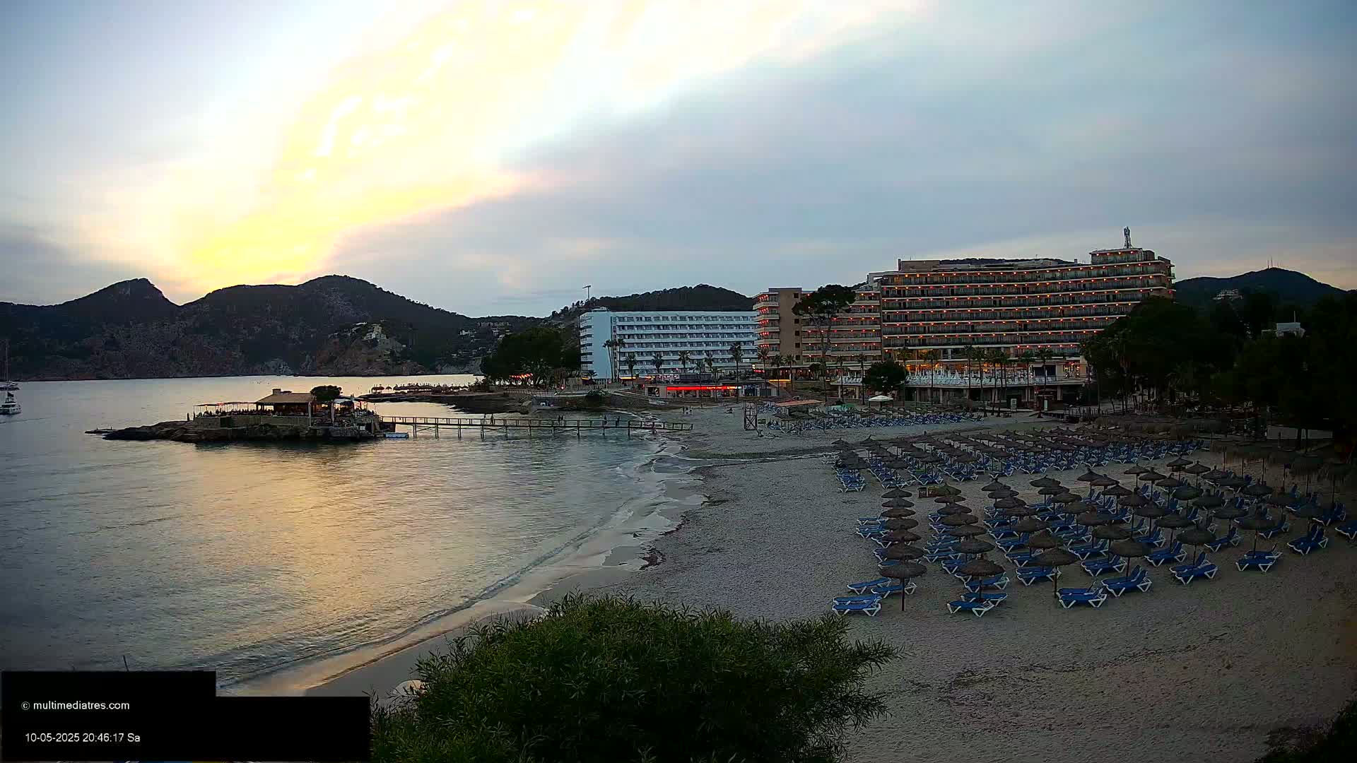 A sandy beach with rows of closed beach umbrellas and lounge chairs sits in front of a large hotel complex at sunset, with calm water and mountains in the background.