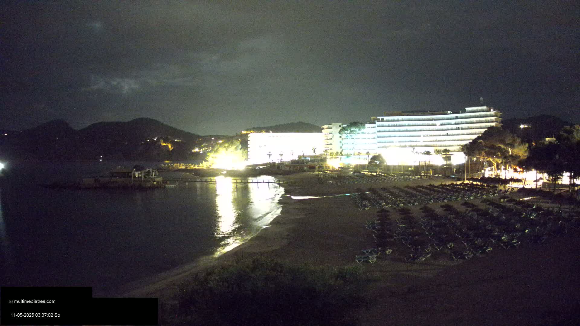 A dark, nighttime scene shows a sandy beach with rows of lounge chairs next to a brightly lit hotel, all set against a backdrop of dark hills and a calm body of water.