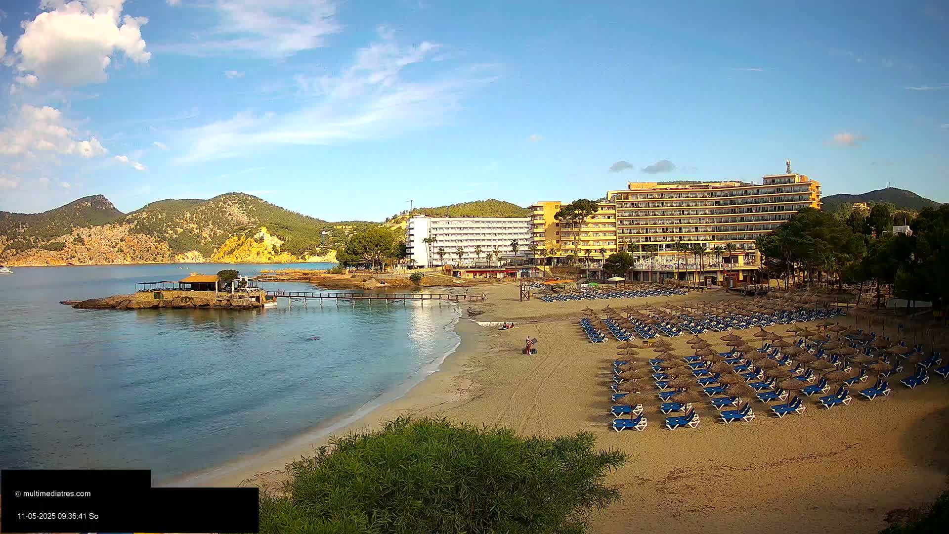 A sandy beach with numerous lounge chairs and umbrellas sits in front of a large hotel complex, backed by green hills under a mostly sunny sky.