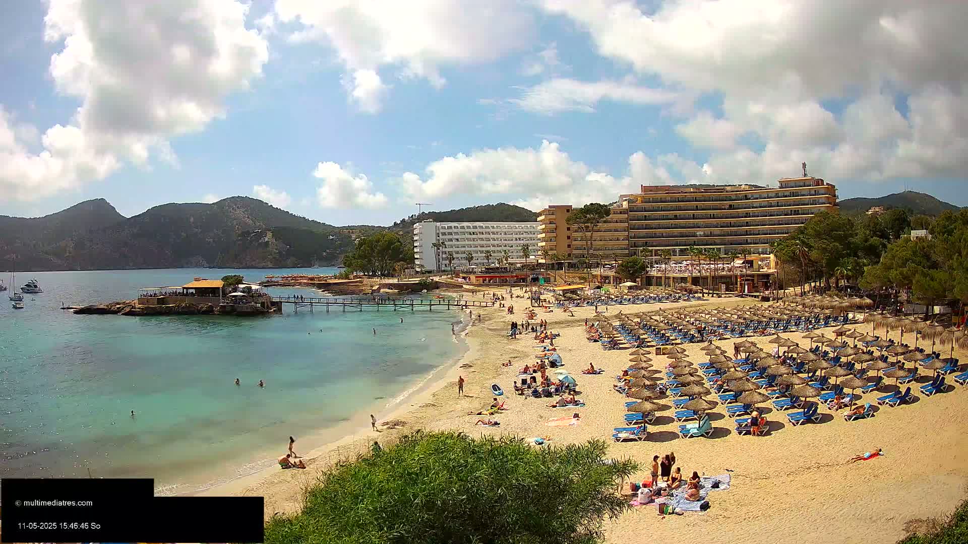 A sandy beach with numerous umbrellas and lounge chairs, a small restaurant on an islet, a large hotel, and calm, clear turquoise water under a partly cloudy sky.