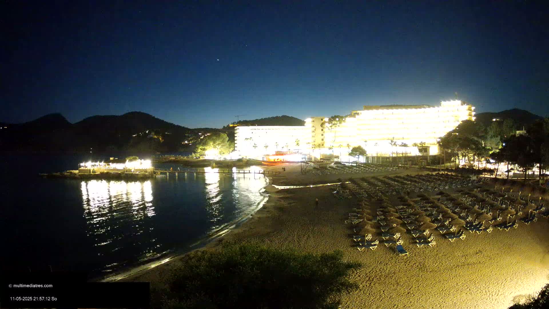 A brightly lit hotel overlooks a sandy beach with rows of lounge chairs at night under a clear, starry sky.