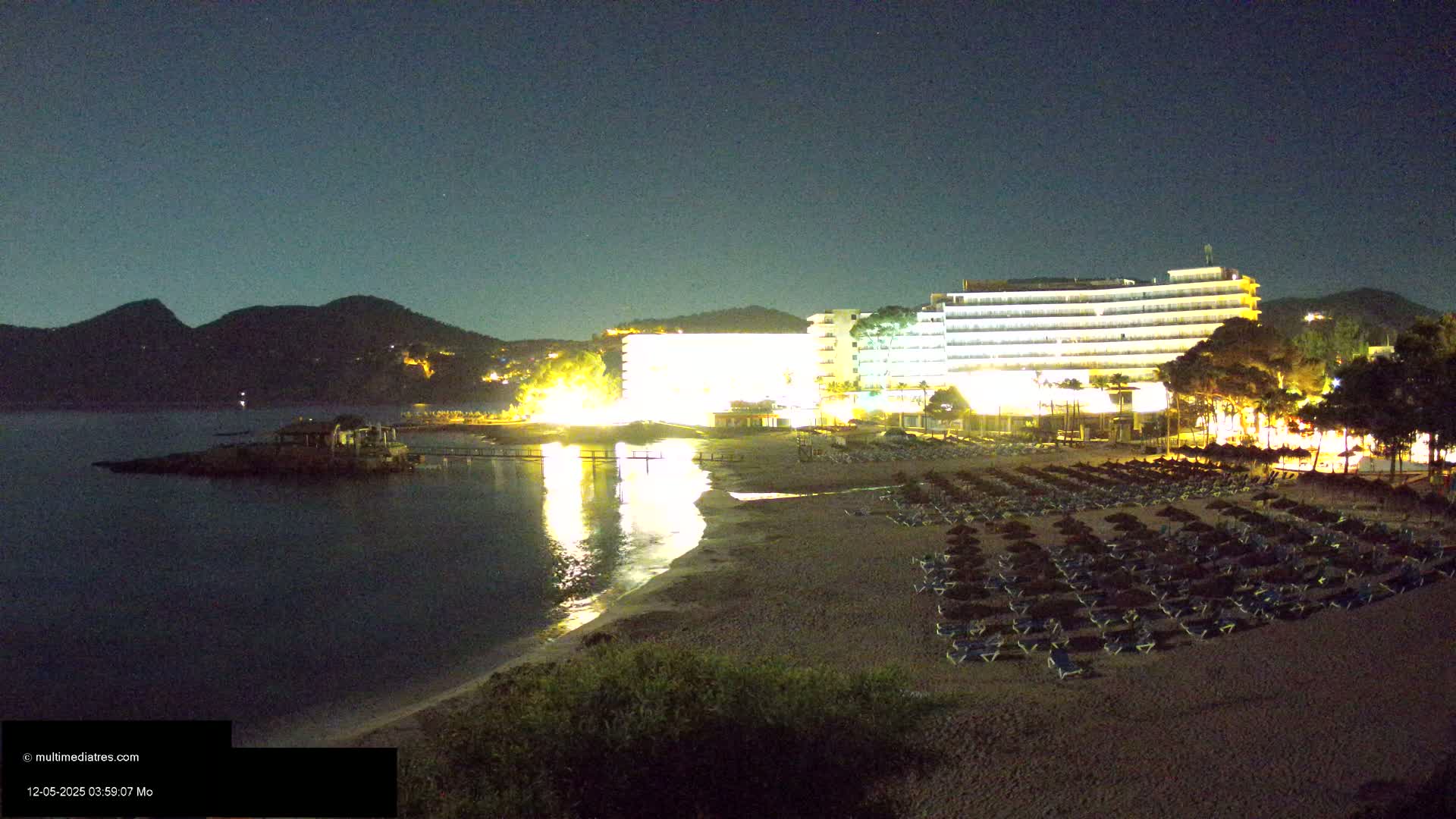 A nighttime view of a sandy beach with rows of lounge chairs and umbrellas in front of a brightly lit hotel, set against a dark, mountainous backdrop under a clear sky.