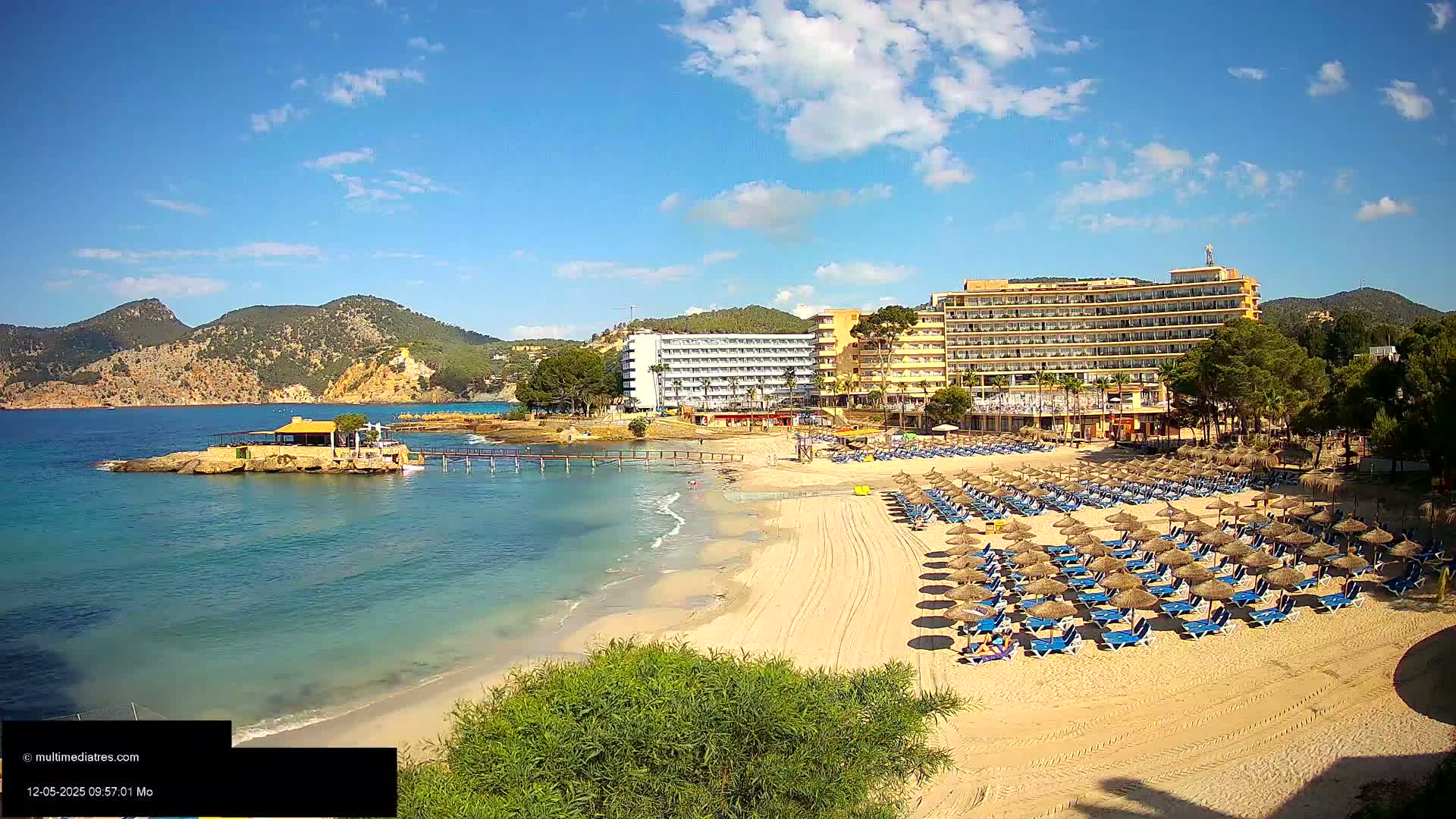 A sandy beach with numerous thatched umbrellas and lounge chairs sits in front of two large hotels, backed by green hills under a mostly sunny sky.