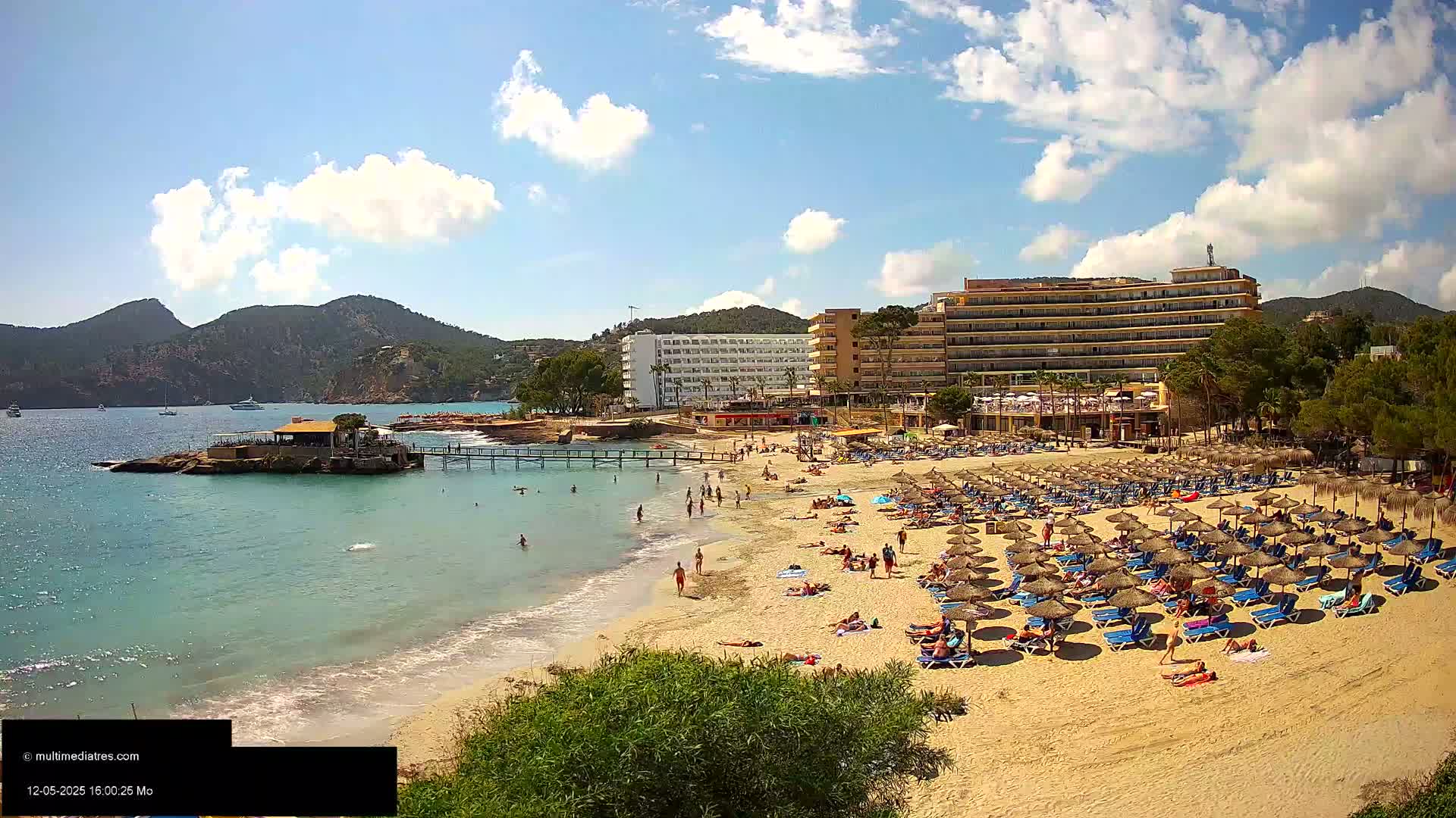 A sandy beach with numerous sunbathers and beach umbrellas is situated in front of a large hotel complex under a partly cloudy sky.