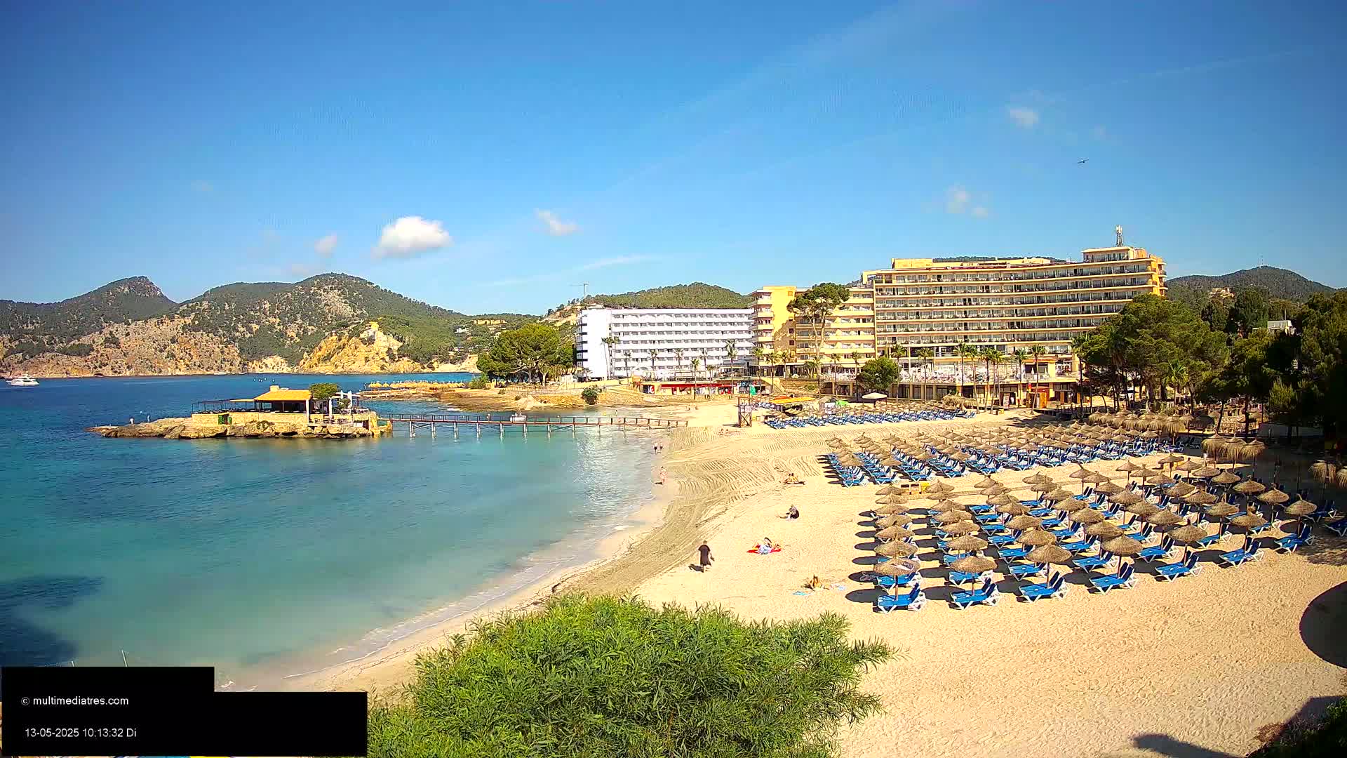 A sandy beach with numerous blue lounge chairs and thatched umbrellas sits in front of a large hotel complex on a clear, sunny day, with calm turquoise water and hills in the background.