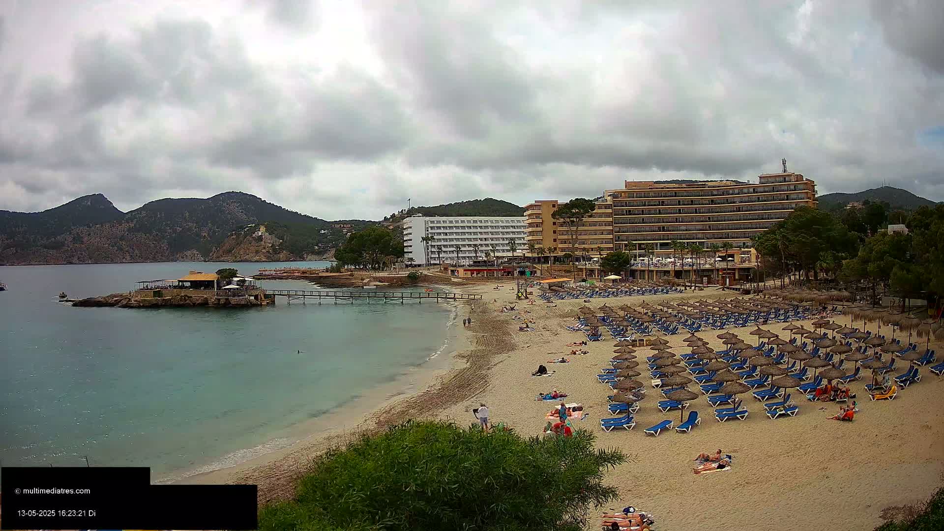 A sandy beach with numerous thatched umbrellas and lounge chairs sits below several hotels, all under a cloudy sky, near a calm, turquoise body of water and rocky coastline.