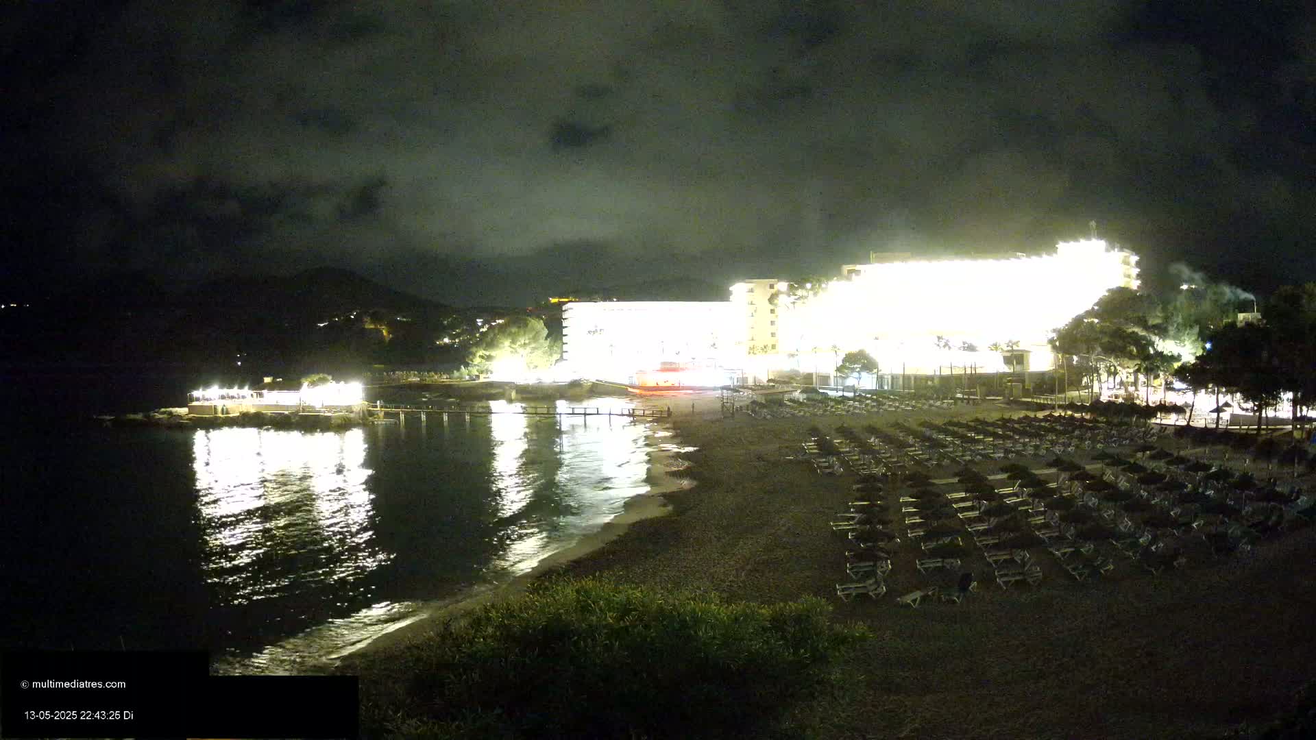 A brightly lit hotel overlooks a dark beach with rows of empty lounge chairs under a nighttime sky.
