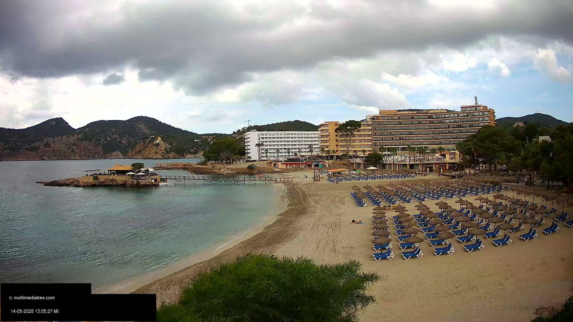 A sandy beach with numerous lounge chairs and umbrellas sits in front of several hotels, backed by hills under a cloudy sky.