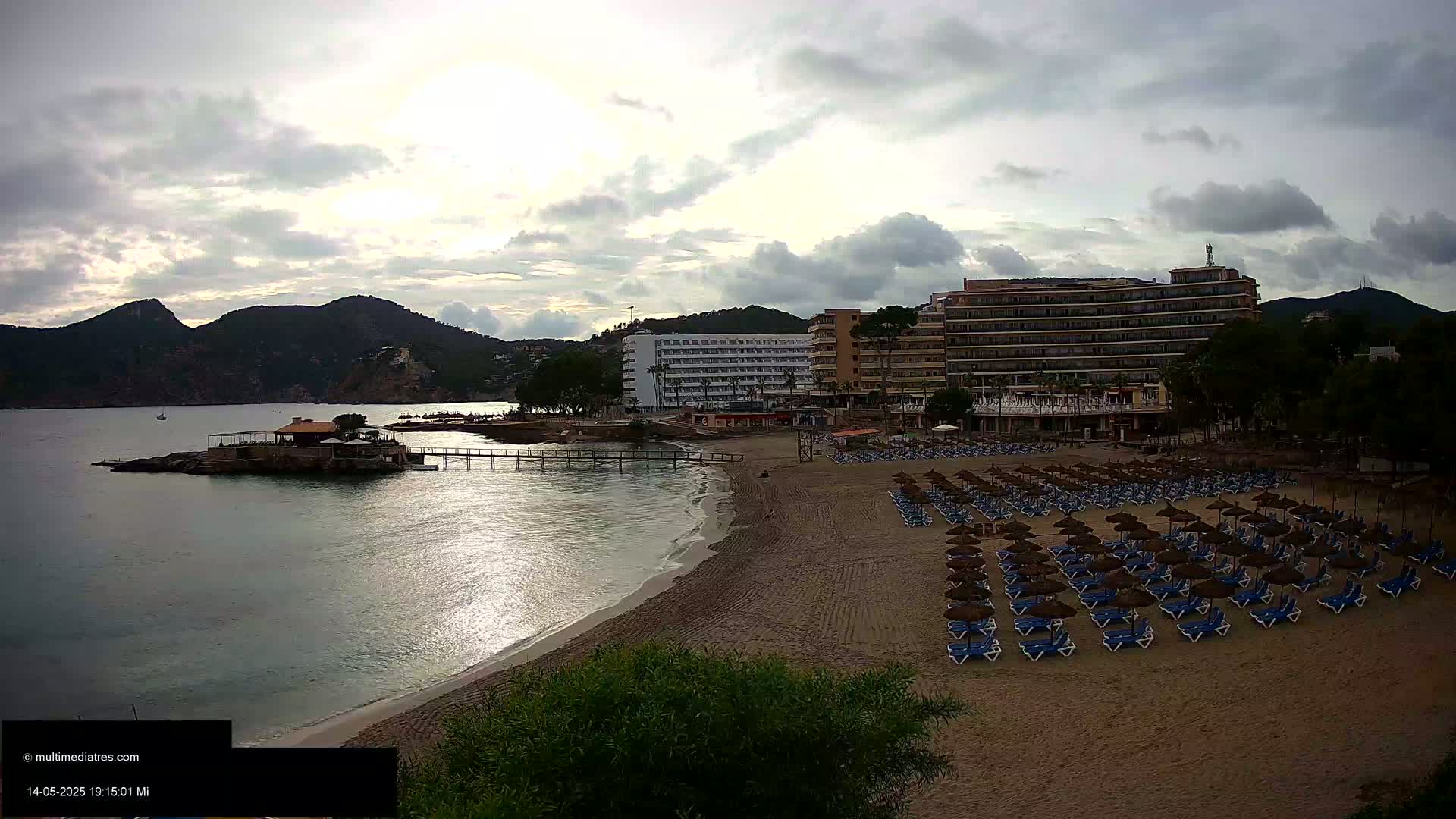 A sandy beach with numerous empty lounge chairs under thatched umbrellas sits in front of hotels and a calm bay under an overcast sky.