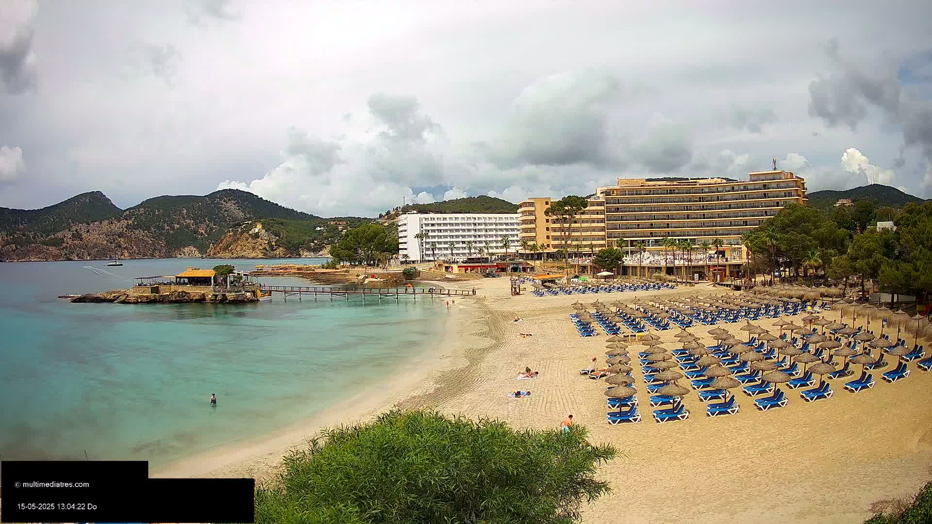 A sandy beach with numerous blue lounge chairs and straw umbrellas sits in front of several hotels under a partly cloudy sky, next to a calm, clear turquoise body of water.
