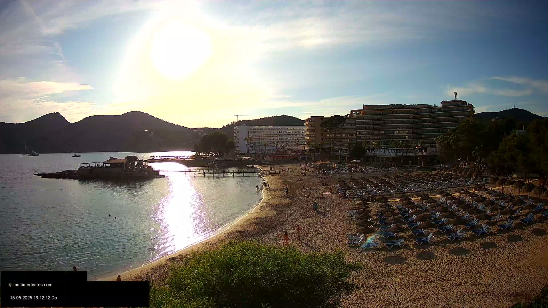 A sandy beach with numerous beach umbrellas and lounge chairs sits below hotels and mountains, under a bright, sunny sky.