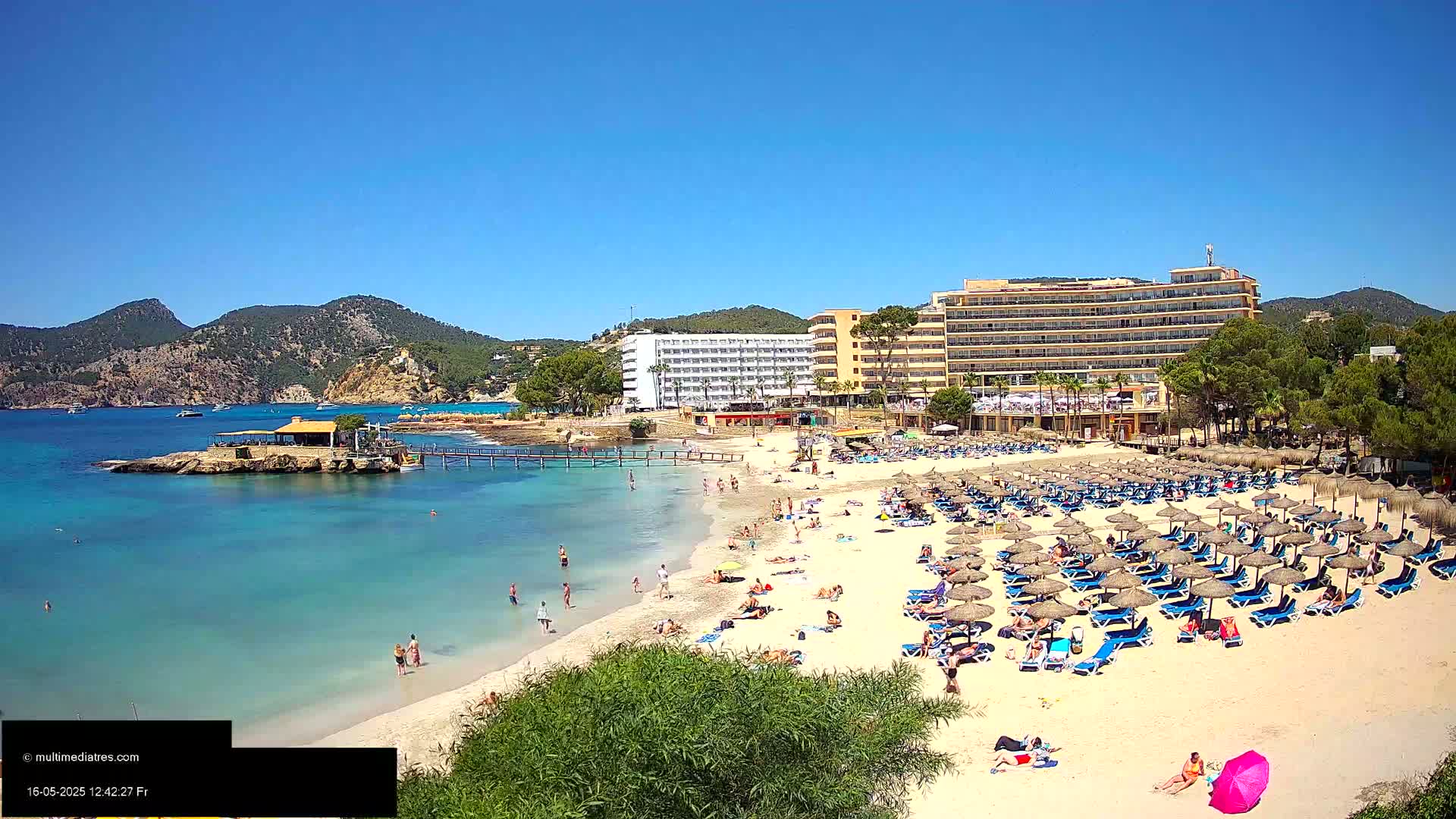 A sandy beach with numerous beach umbrellas and lounge chairs, many people sunbathing and swimming, sits in front of a large hotel complex, all under a clear blue sky.
