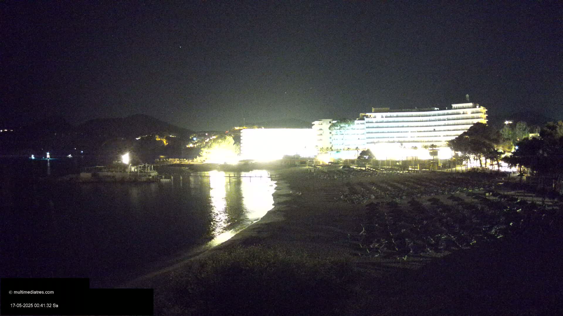 A brightly lit hotel is situated on a dark beach at night, with calm water reflecting the lights.