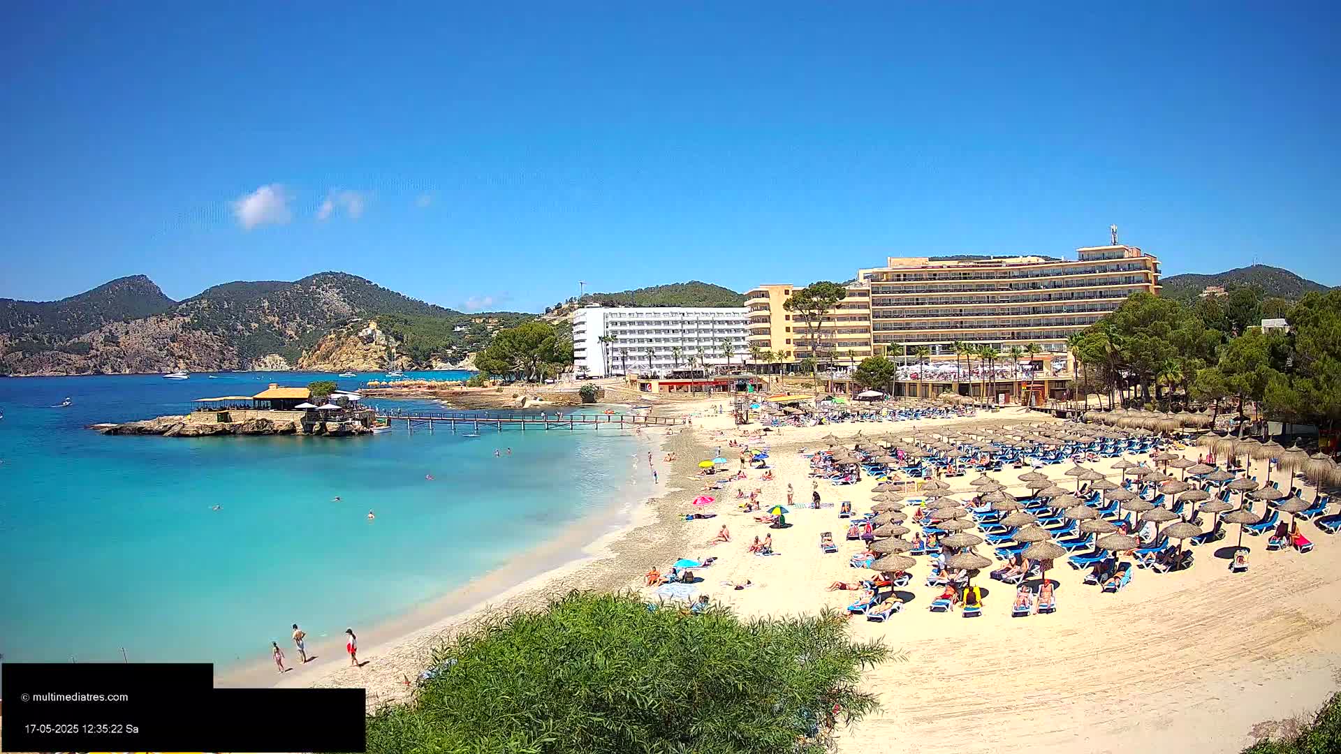 A sandy beach with numerous thatched umbrellas and sunbathers is situated in front of a large hotel complex, backed by green hills under a clear blue sky.