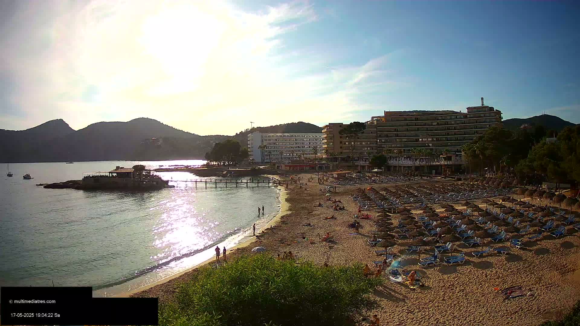 A sandy beach with numerous lounge chairs and umbrellas sits below several hotels, near a calm bay under a mostly sunny sky.