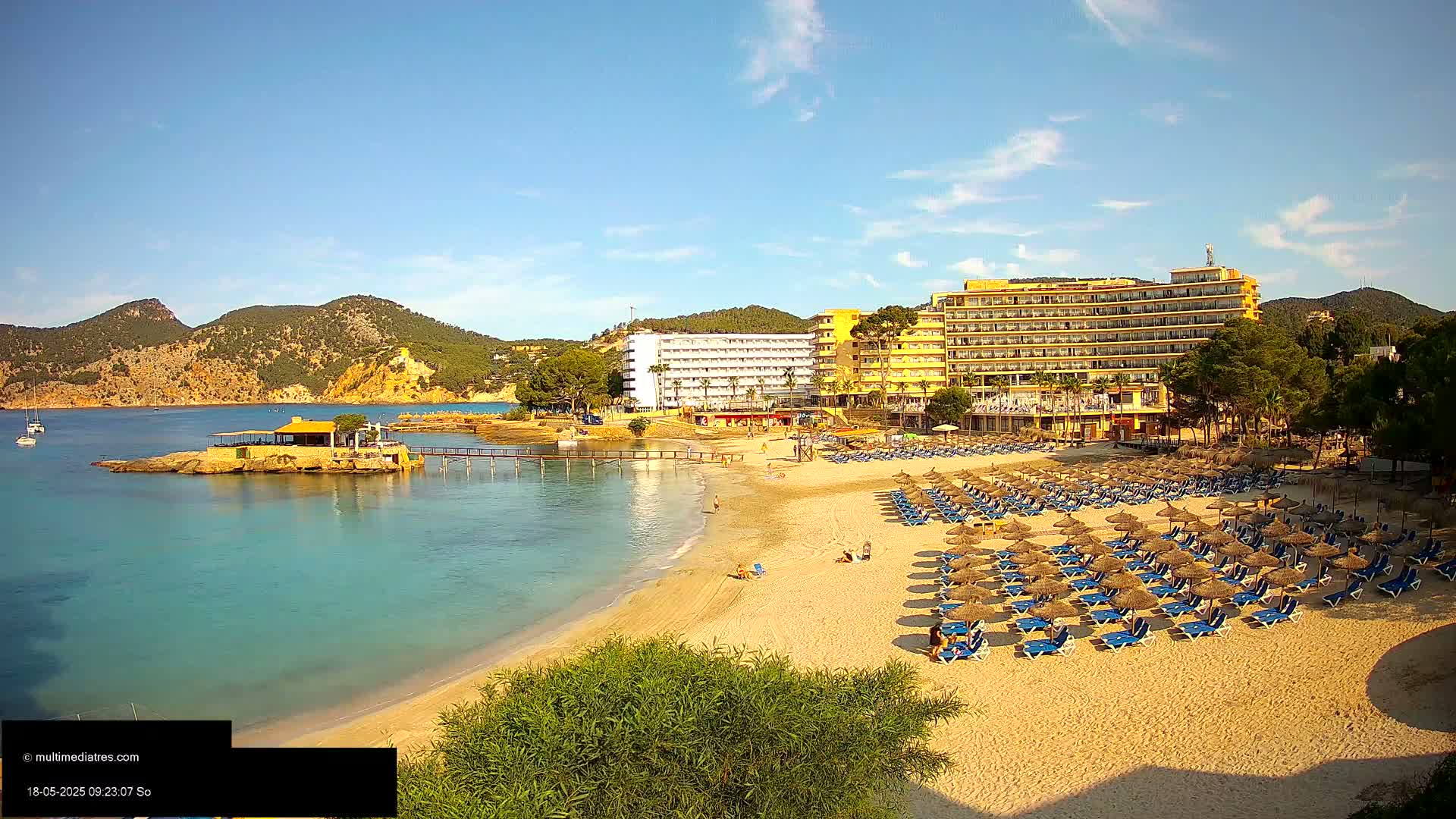 A sandy beach with numerous blue lounge chairs and thatched umbrellas sits in front of a large yellow hotel, all under a mostly clear, sunny sky, with calm turquoise water and hills in the background.