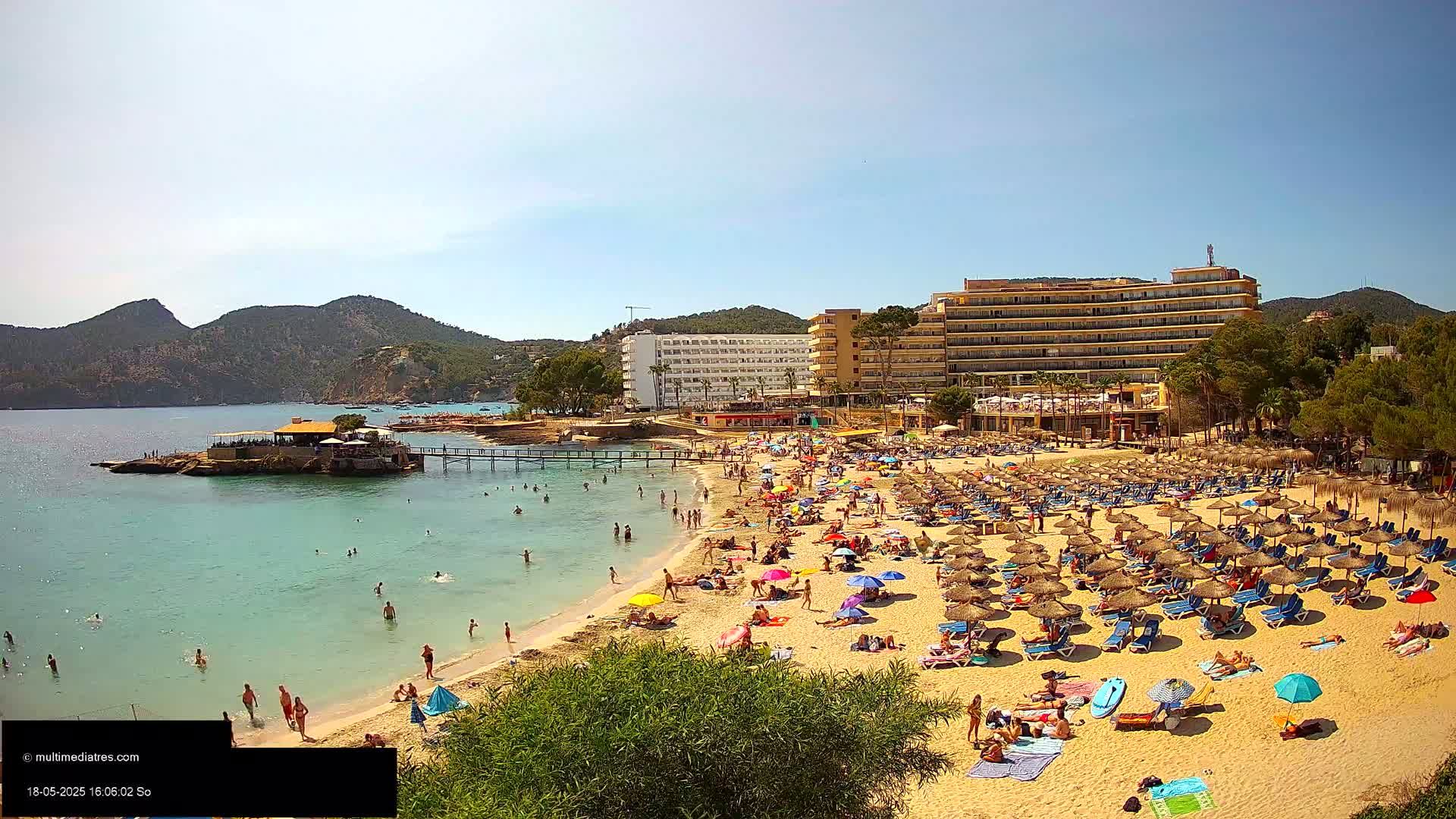 A sunny beach scene shows numerous people relaxing under umbrellas and in lounge chairs near a large hotel and calm turquoise water, backed by lush green hills.