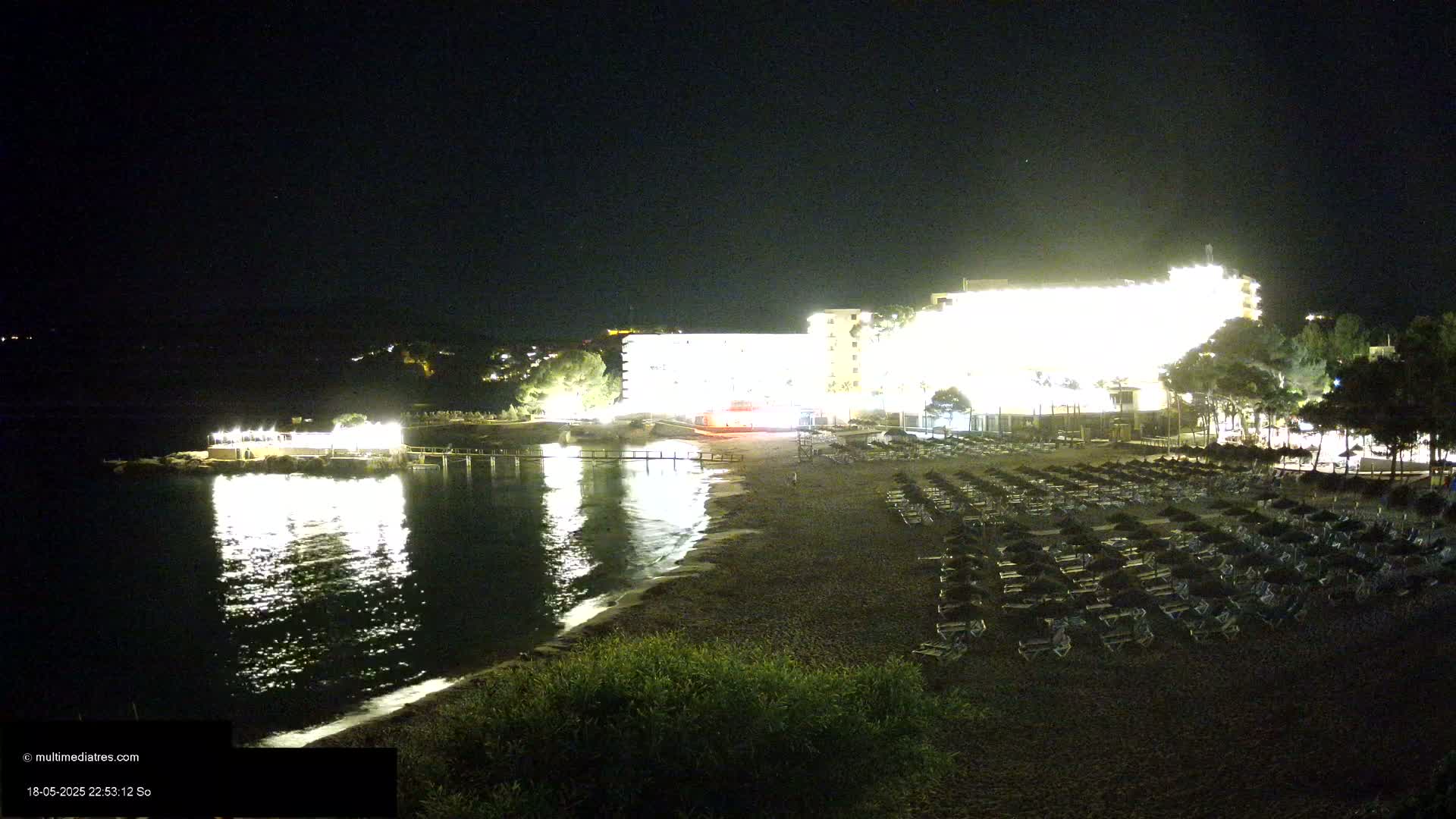 A brightly lit hotel sits on a dark beach at night, next to calm water reflecting the lights.