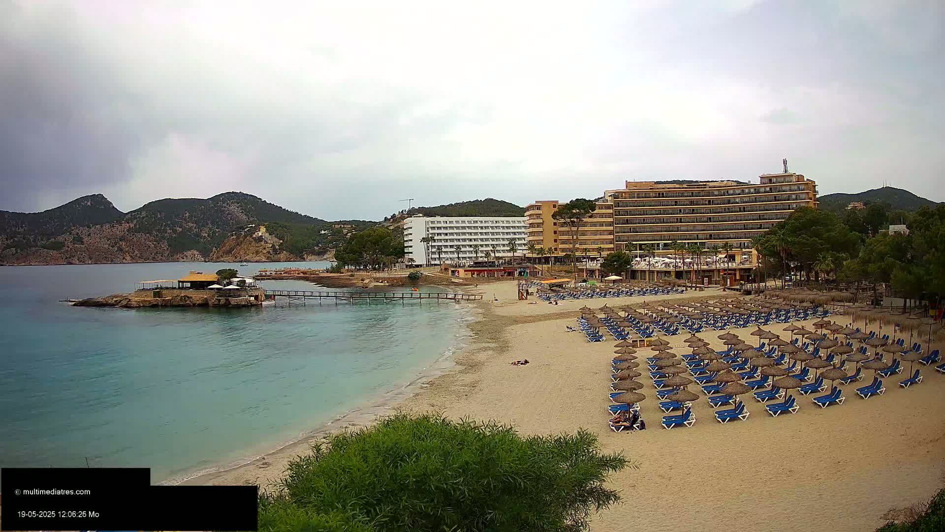A sandy beach with numerous blue lounge chairs under thatched umbrellas sits in front of several hotels, all under an overcast sky, near a calm body of water and green hills.