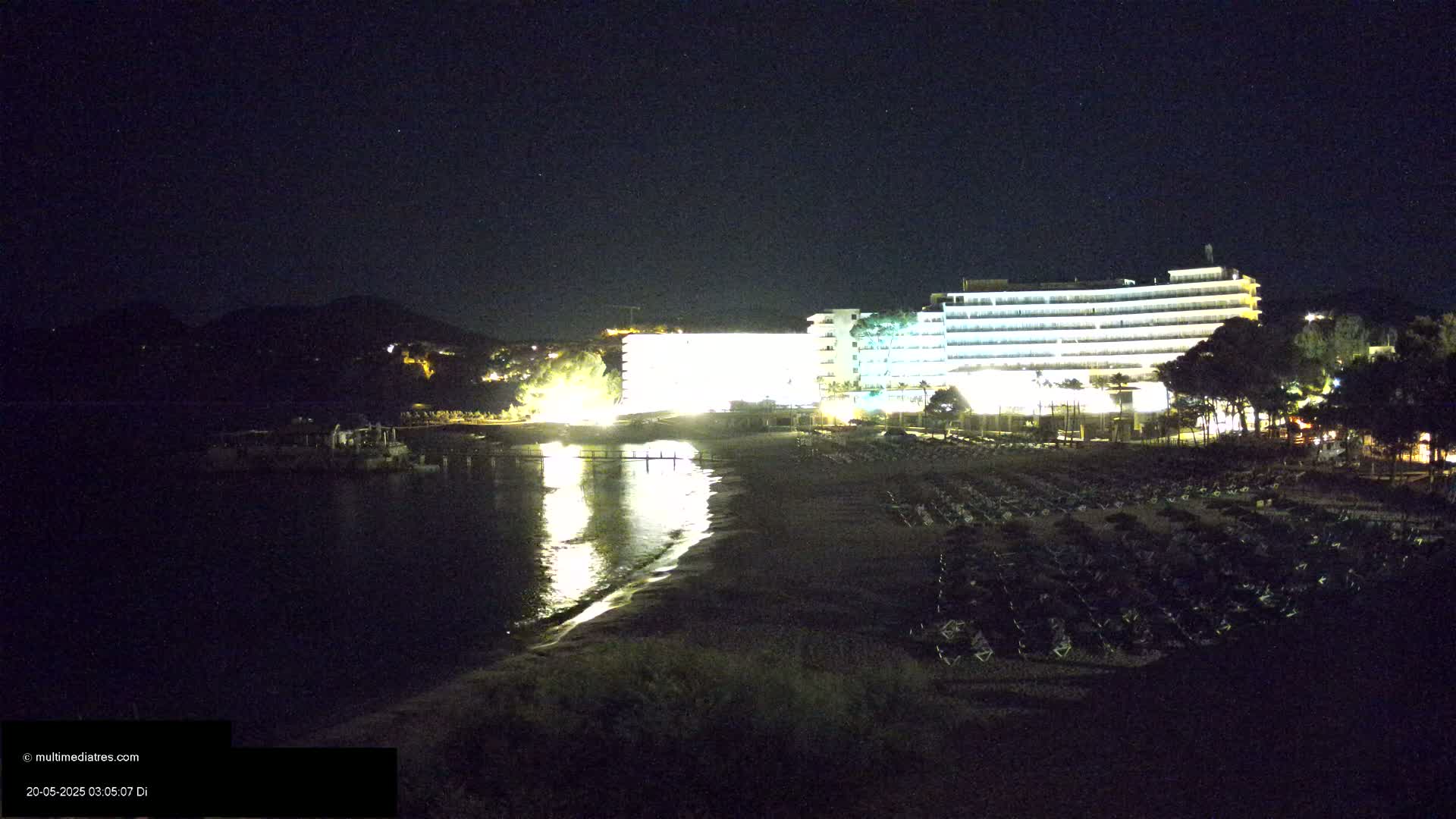 A brightly lit hotel is situated on a dark beach at night, overlooking a calm body of water under a starry sky.