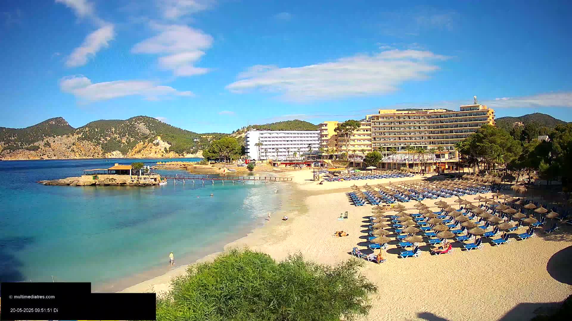 A sandy beach with numerous blue lounge chairs and thatched umbrellas sits in front of a large hotel complex, all under a mostly sunny sky with some scattered clouds, beside a calm, clear turquoise sea.